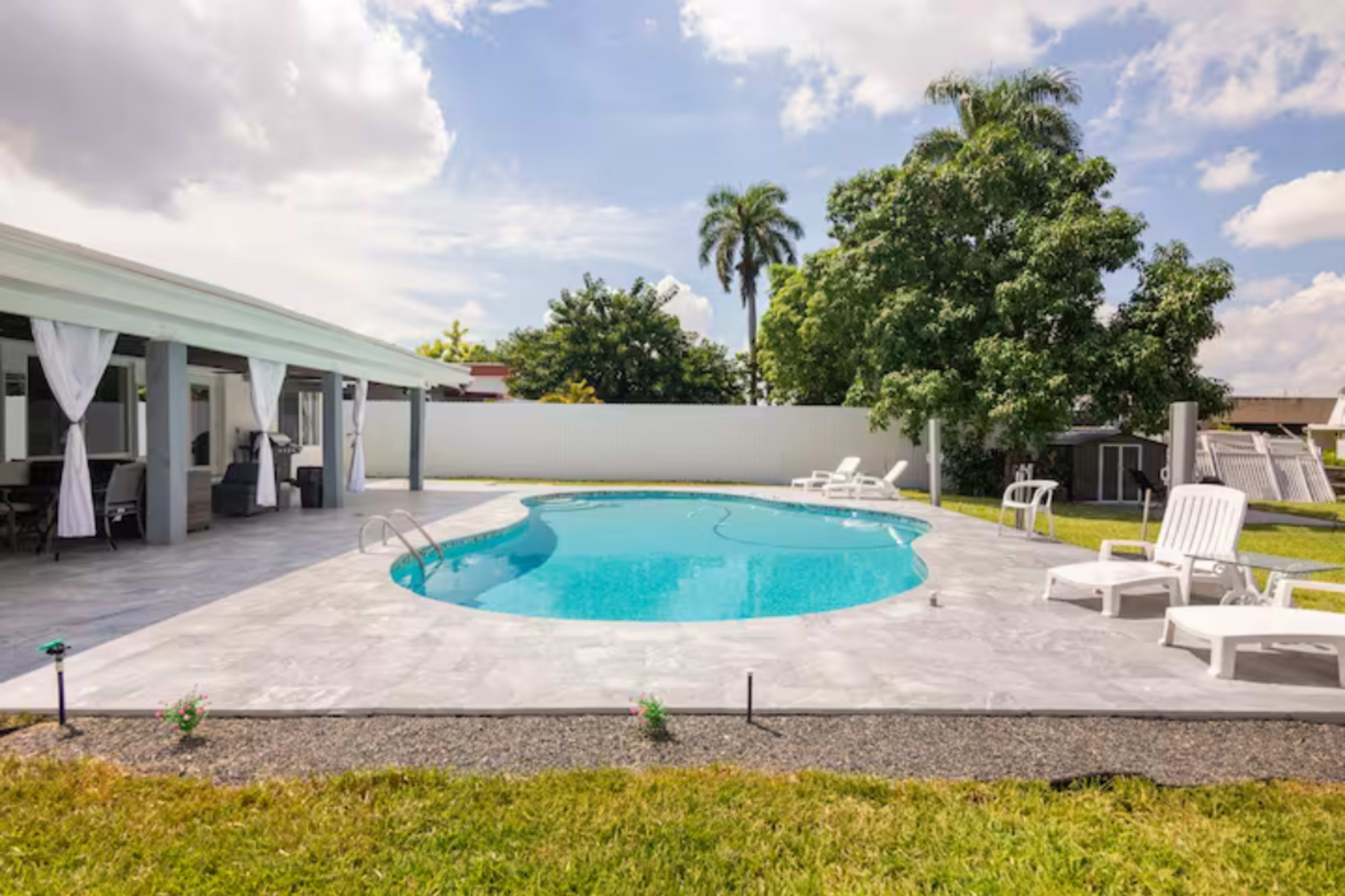 A bright outdoor space features a swimming pool surrounded by lounge chairs and green grass, with a house and palm trees in the background.