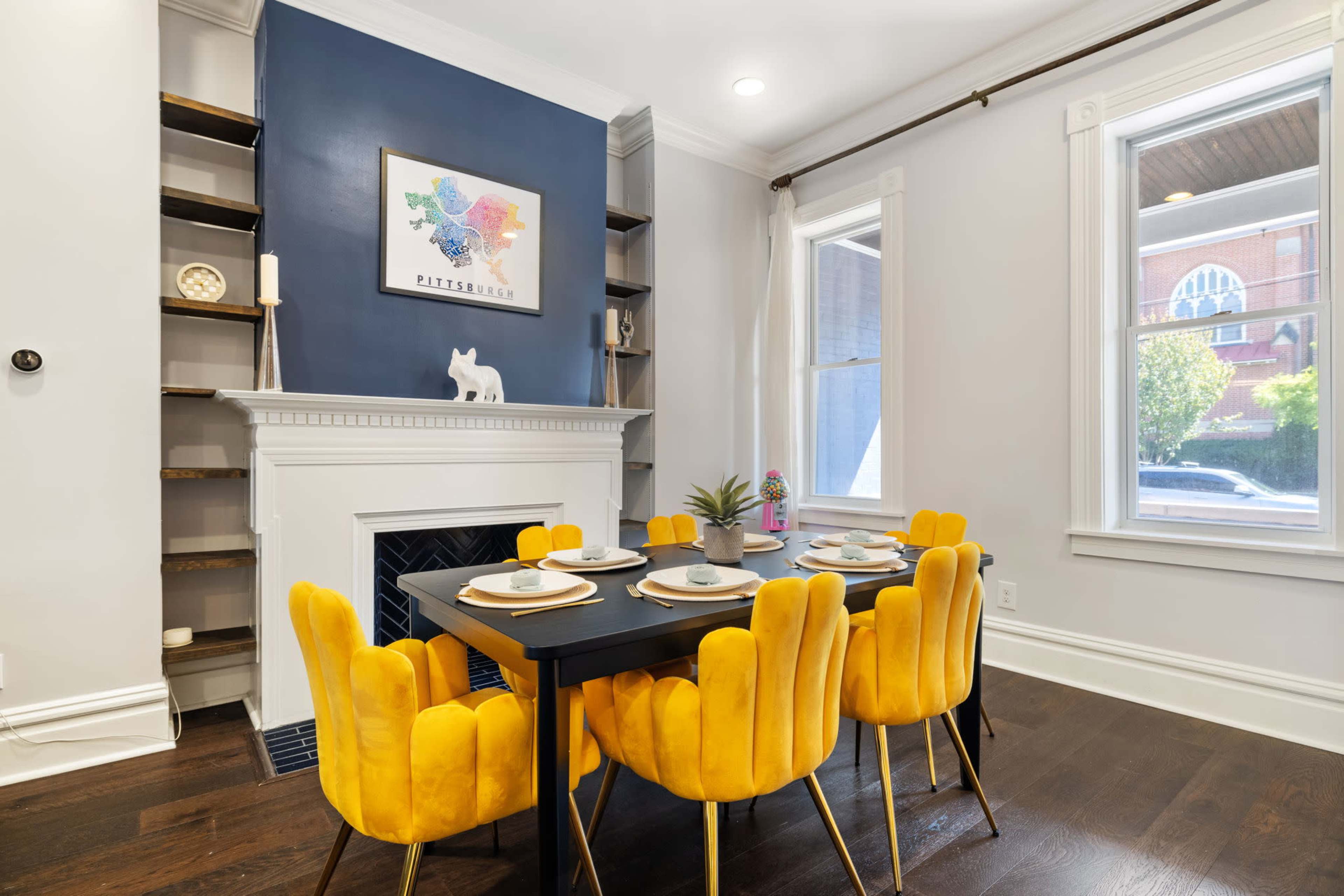 A dining area features a black table surrounded by six yellow chairs, with a colorful art piece above a white fireplace and shelves on the wall.