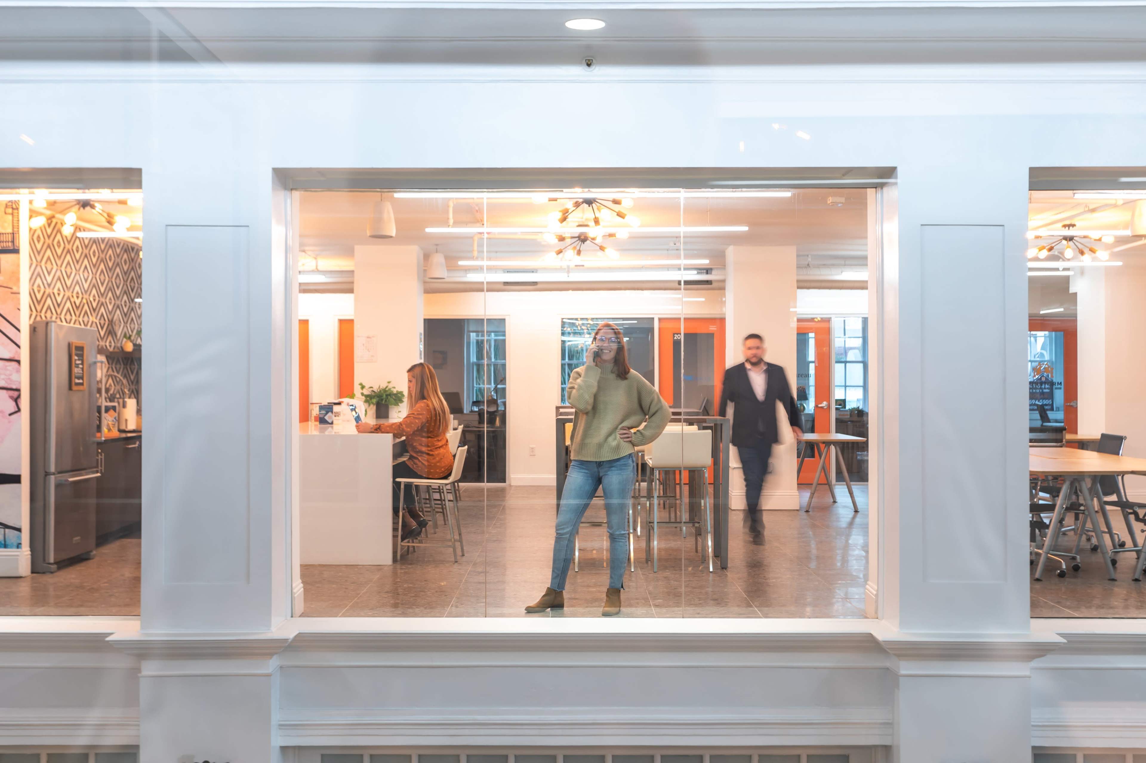 The image depicts an office space with a woman standing confidently in the foreground, while a man walks in the background near a kitchen area where another woman is seated at a table.