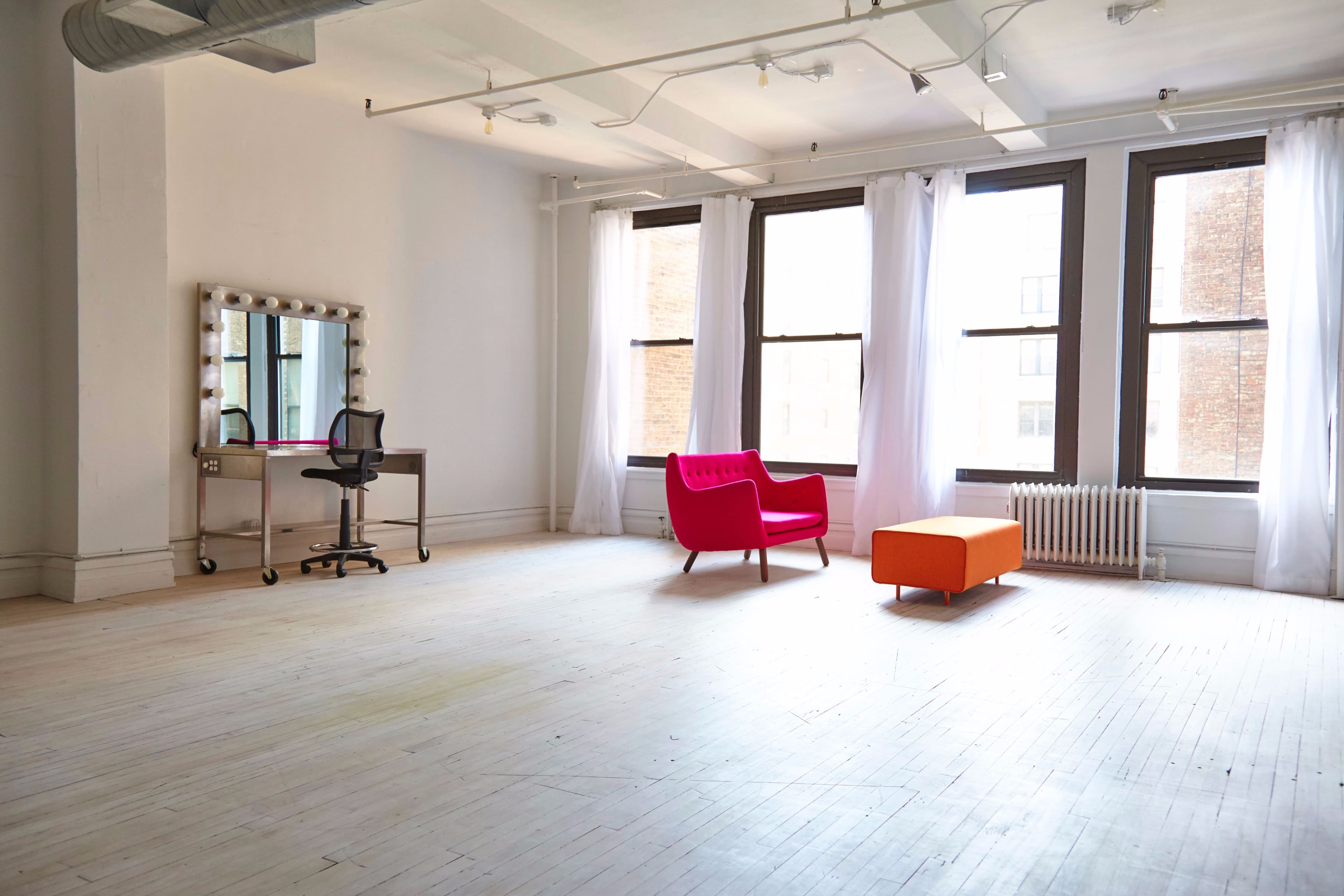 The image shows a spacious, well-lit room with large windows, a pink chair, an orange ottoman, and a vanity mirror with lights.