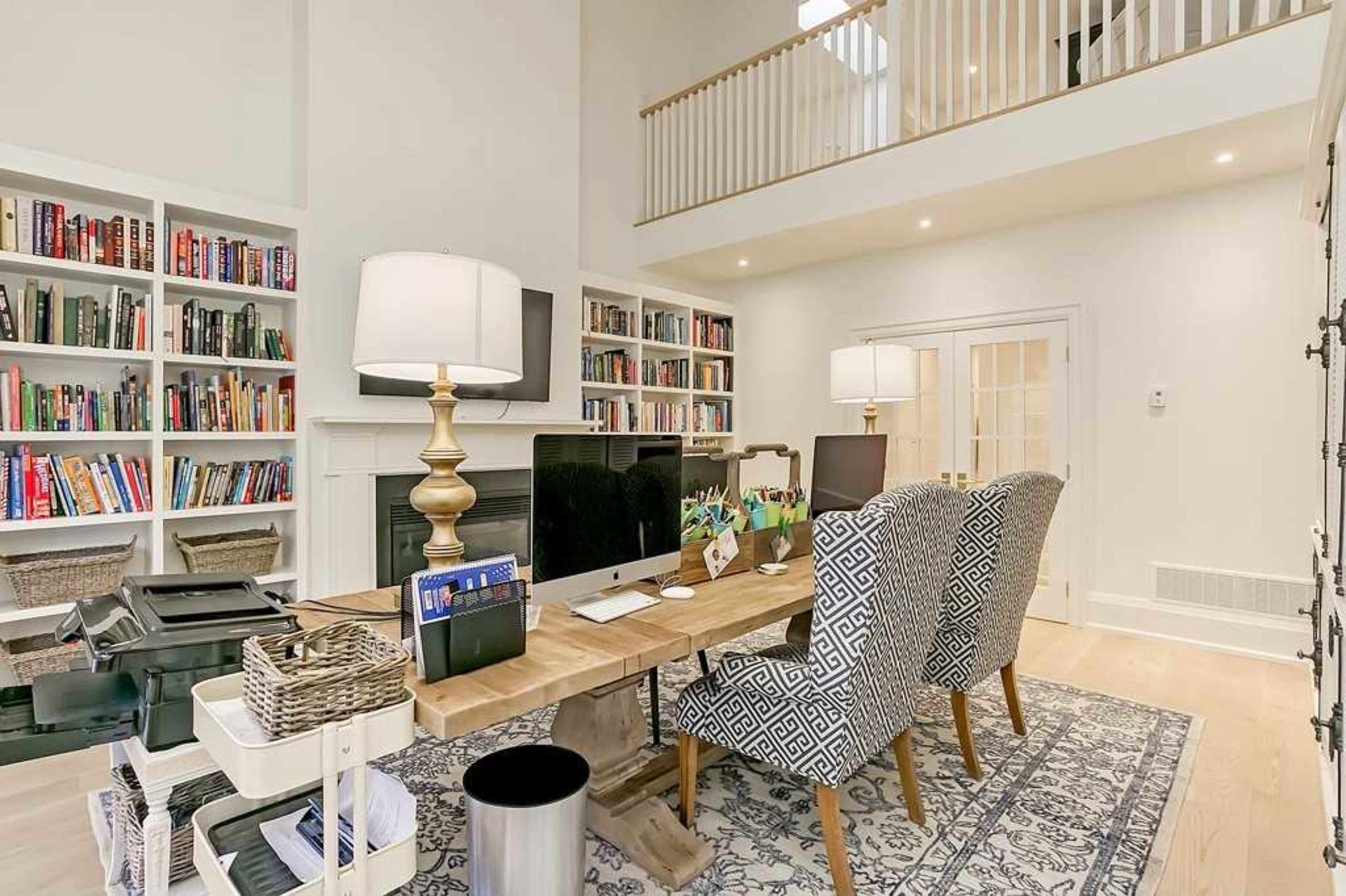 The image shows a modern office space featuring a large wooden desk with two patterned chairs, surrounded by bookshelves filled with books and a staircase leading to a loft area.