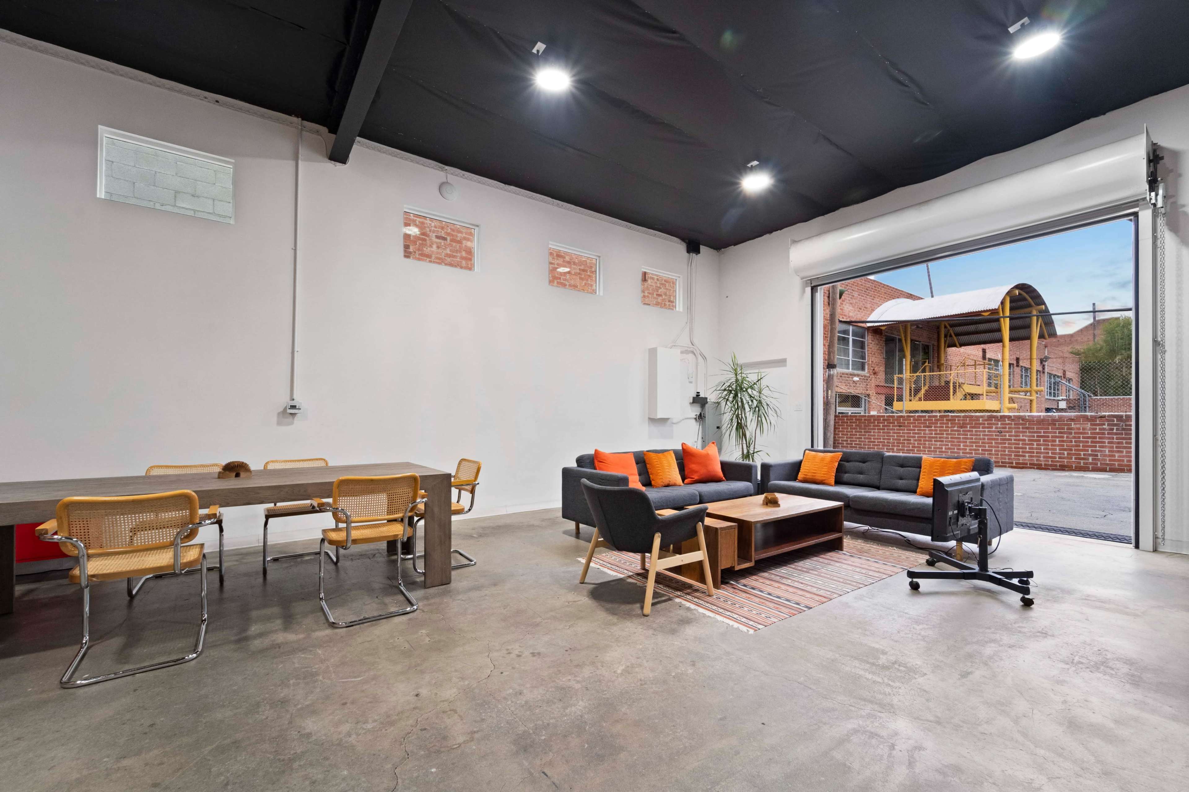 A modern workspace with a seating area featuring a couch and chairs, a dining table, and large windows that provide a view of an outdoor play area.
