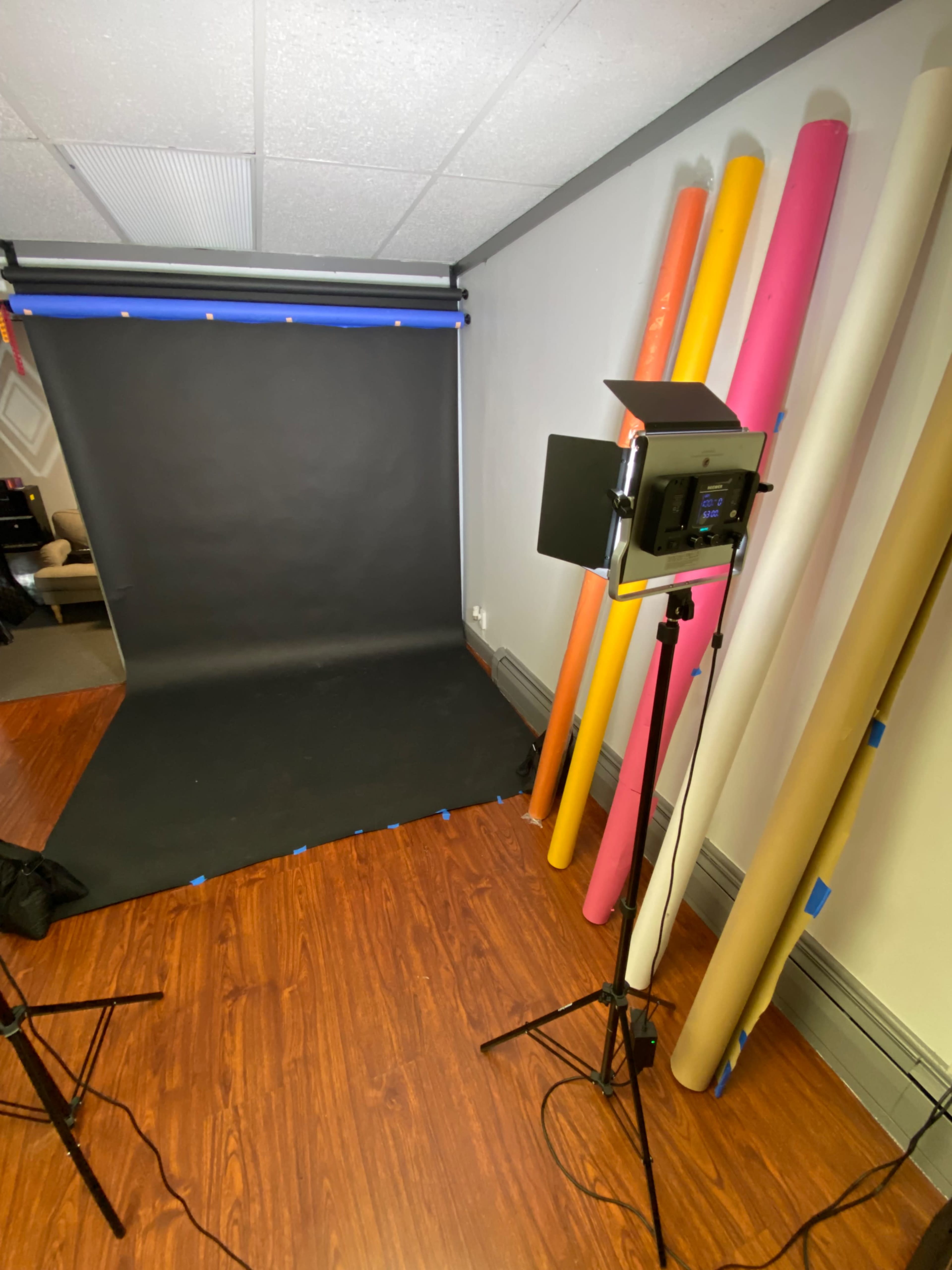 A photography setup featuring a black backdrop, a lighting fixture on a stand, and several rolls of colored paper against the wall.