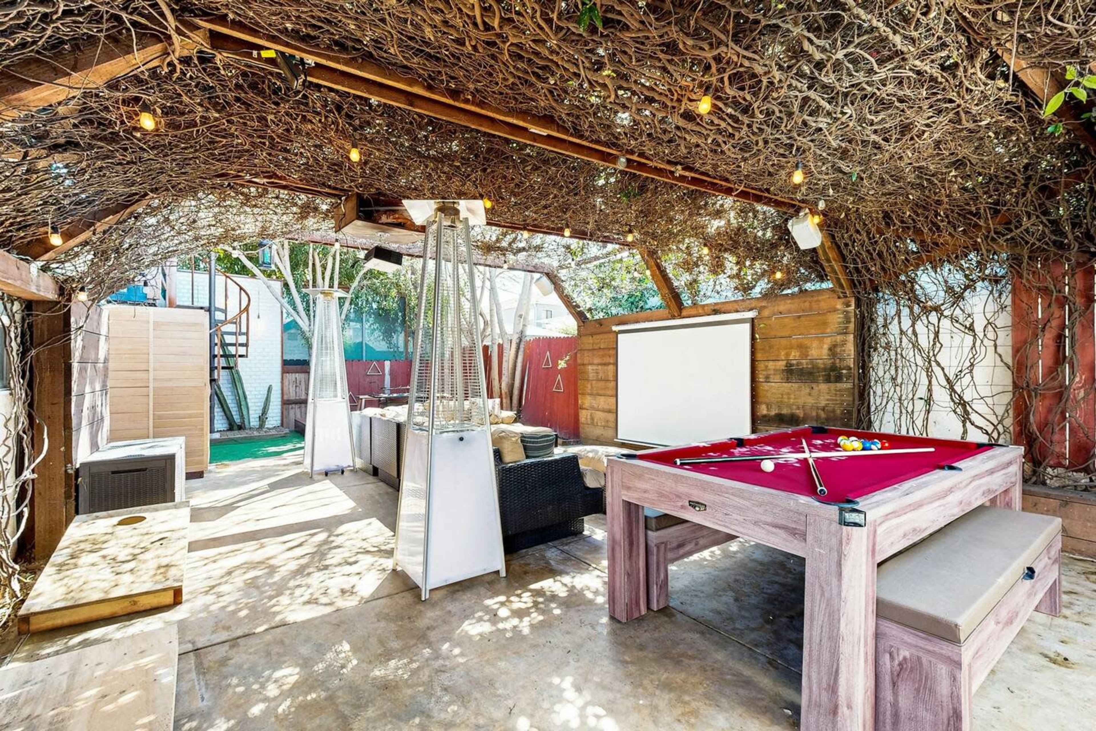 An outdoor entertainment area features a billiards table, a wood-paneled wall, and a canopy of intertwined vines with string lights.