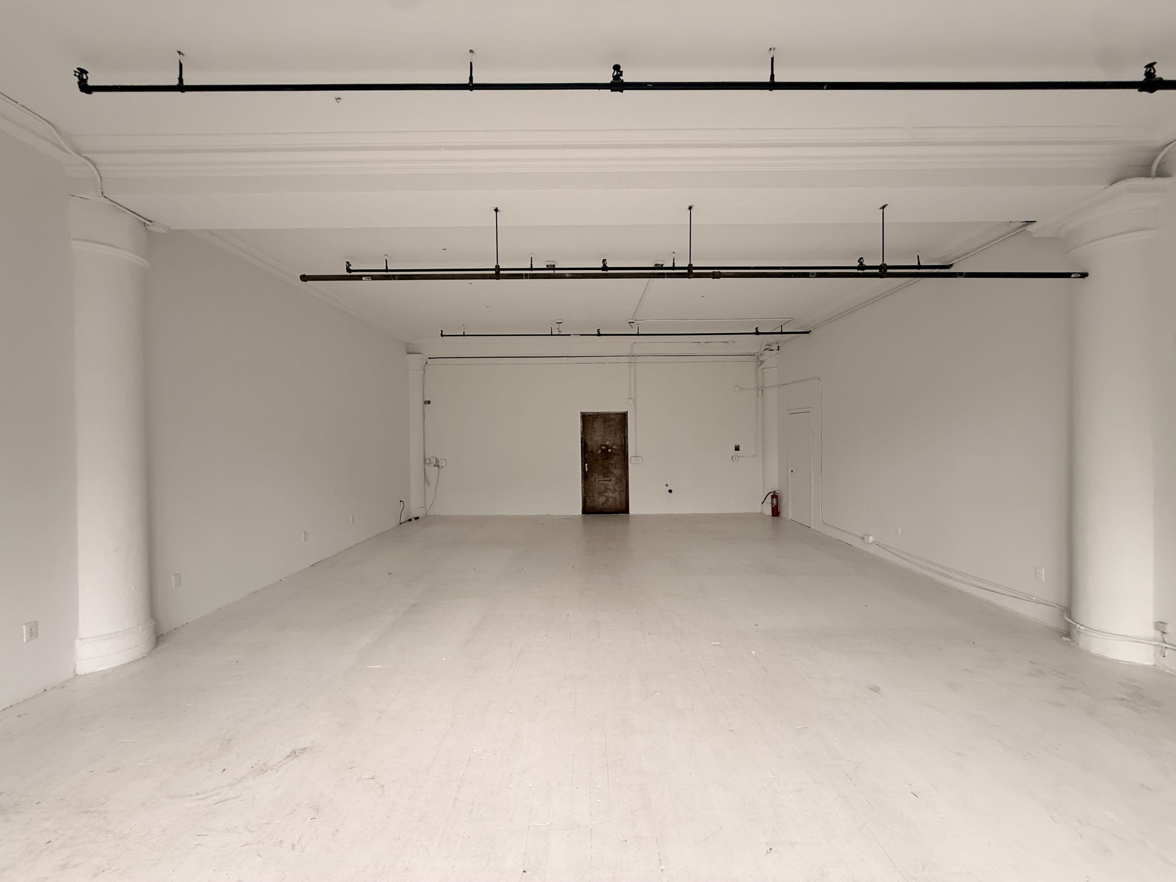 The image shows a spacious, empty room with white walls and a bare wooden door at one end.