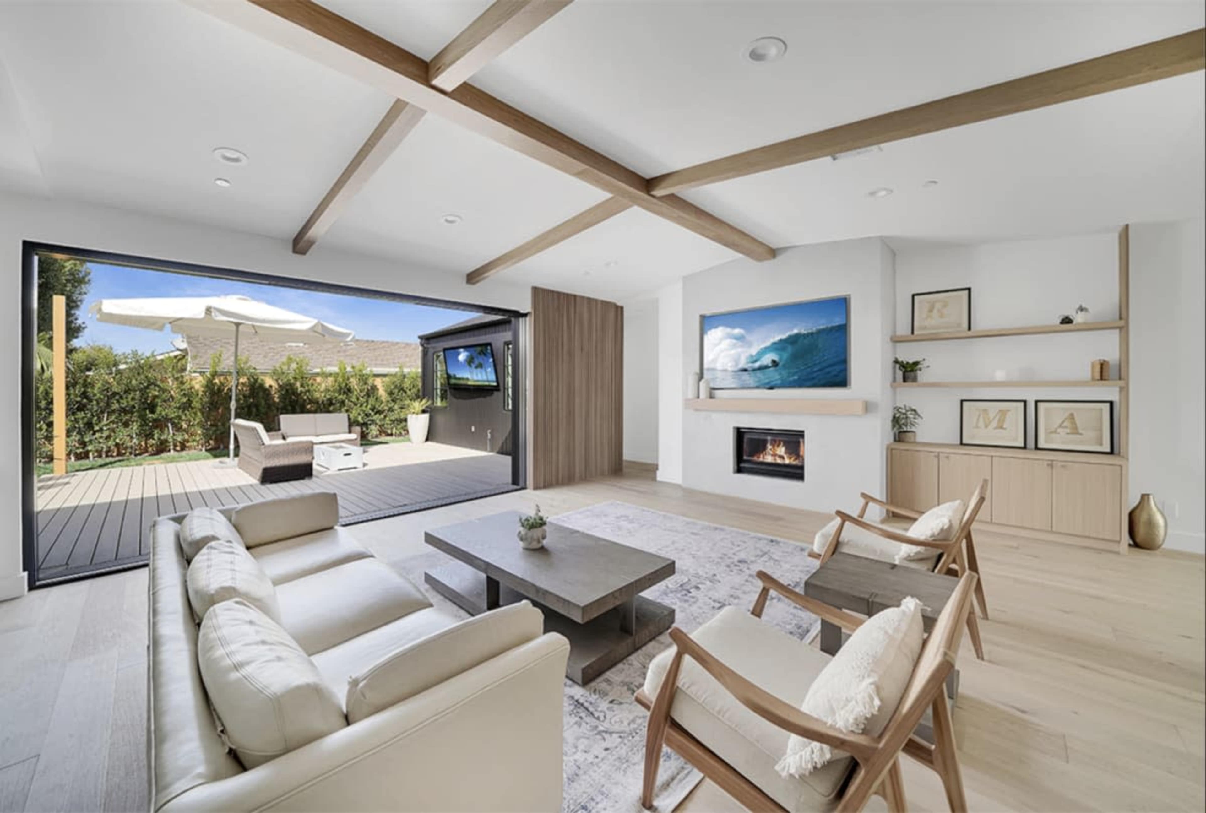 The image shows a modern living room with a fireplace, two armchairs, a beige sofa, and large windows overlooking an outdoor patio area.