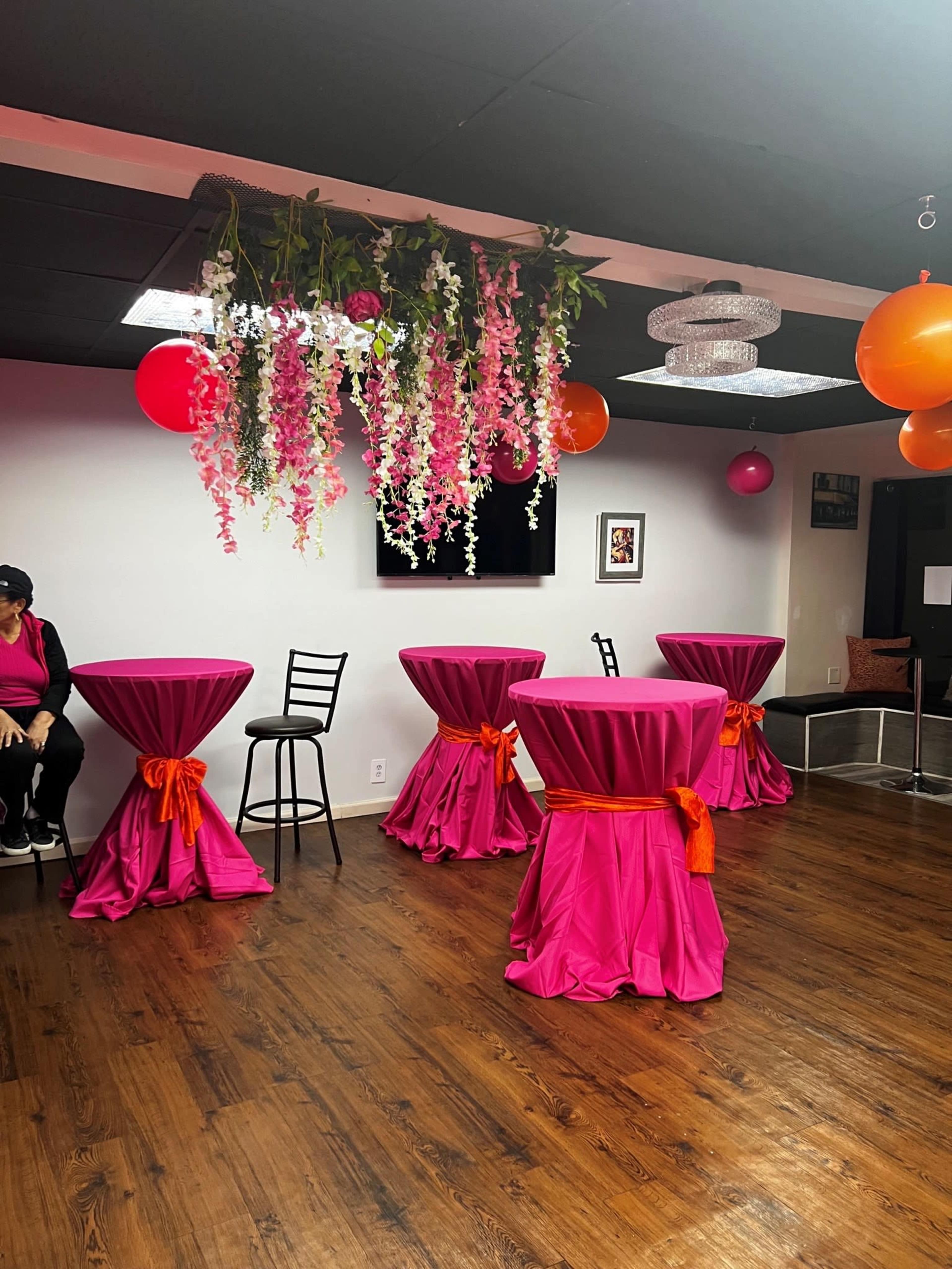 A party room with pink tablecloths adorned with orange sashes, decorative hanging flowers, and colorful balloons.