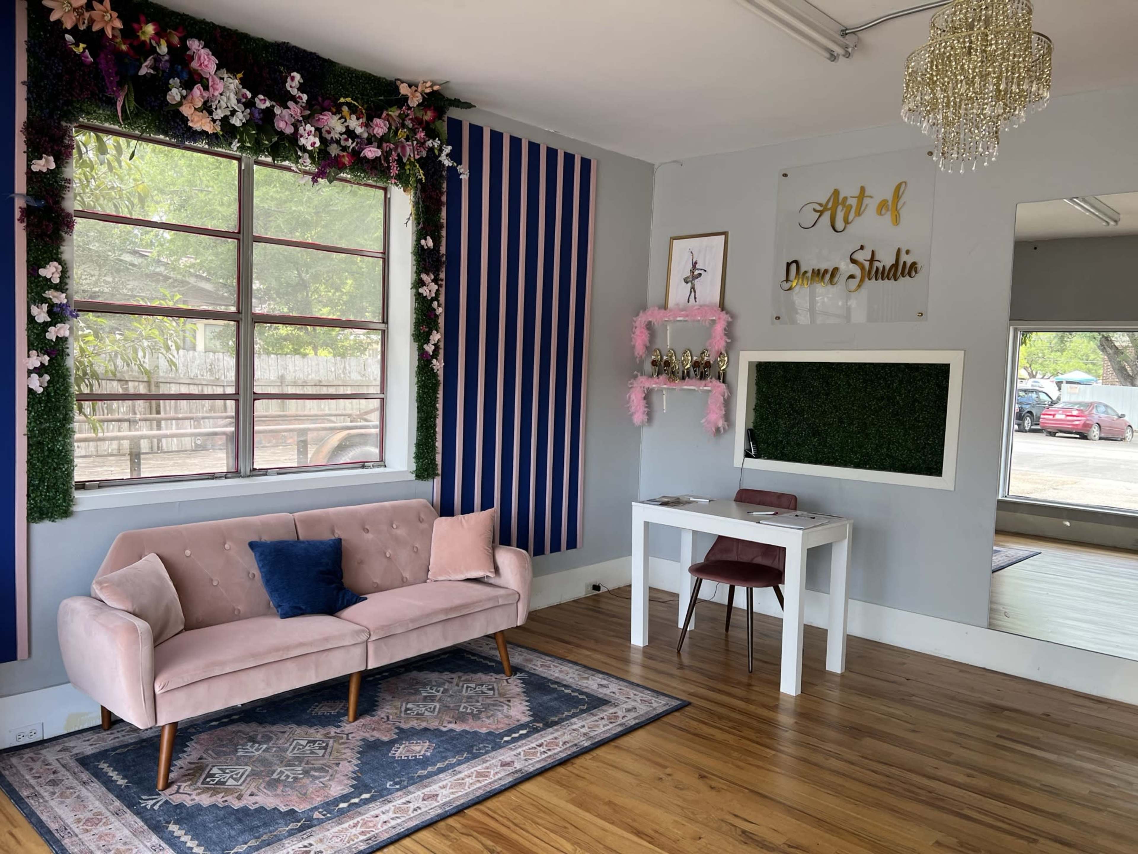 A cozy studio space featuring a pink sofa, a small table with a chair, and decorative elements on the walls, including a floral arrangement and a mirror.