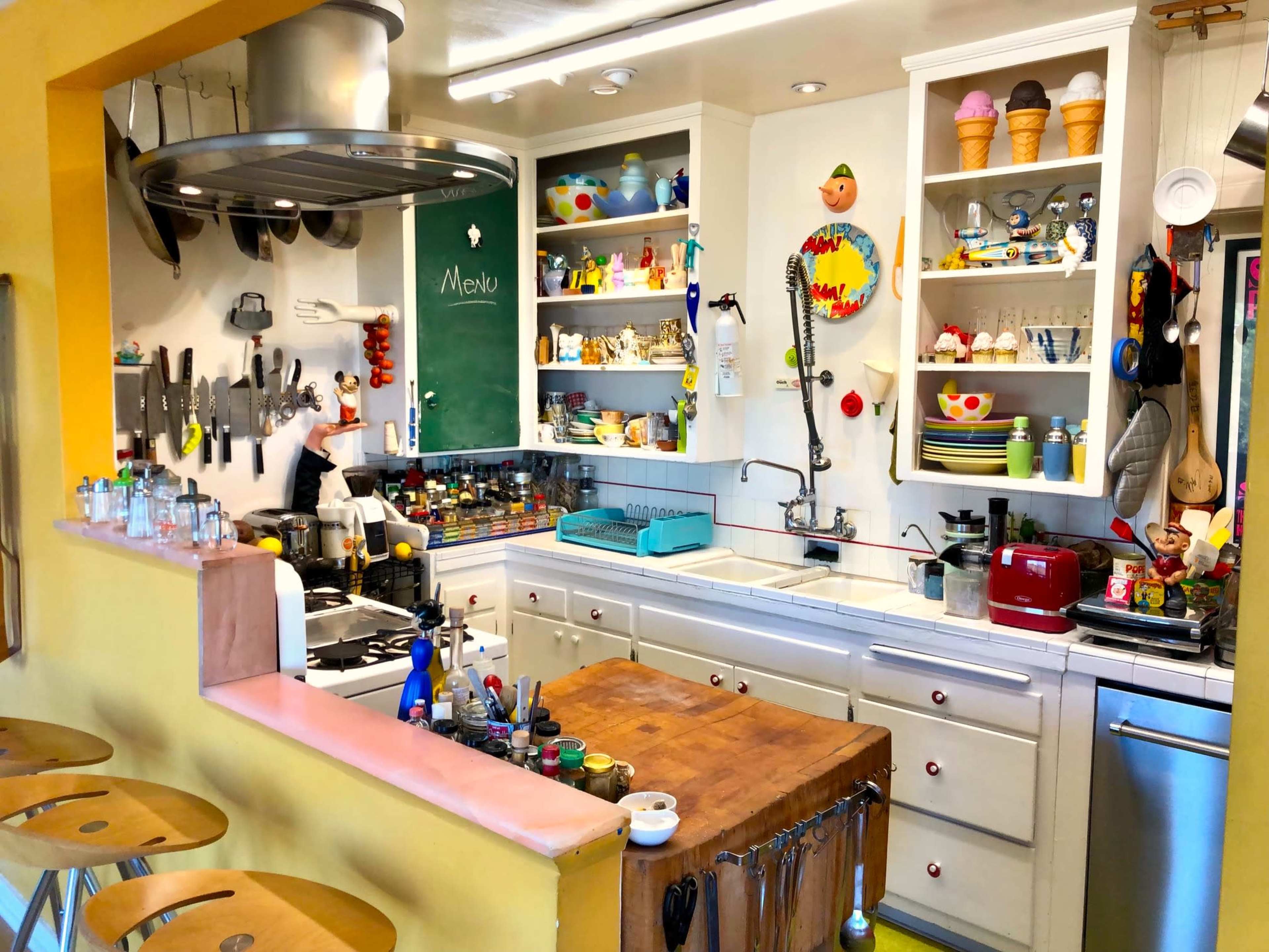 The image shows a colorful kitchen with a wooden island, shelves filled with various kitchenware, and a menu chalkboard on the wall.