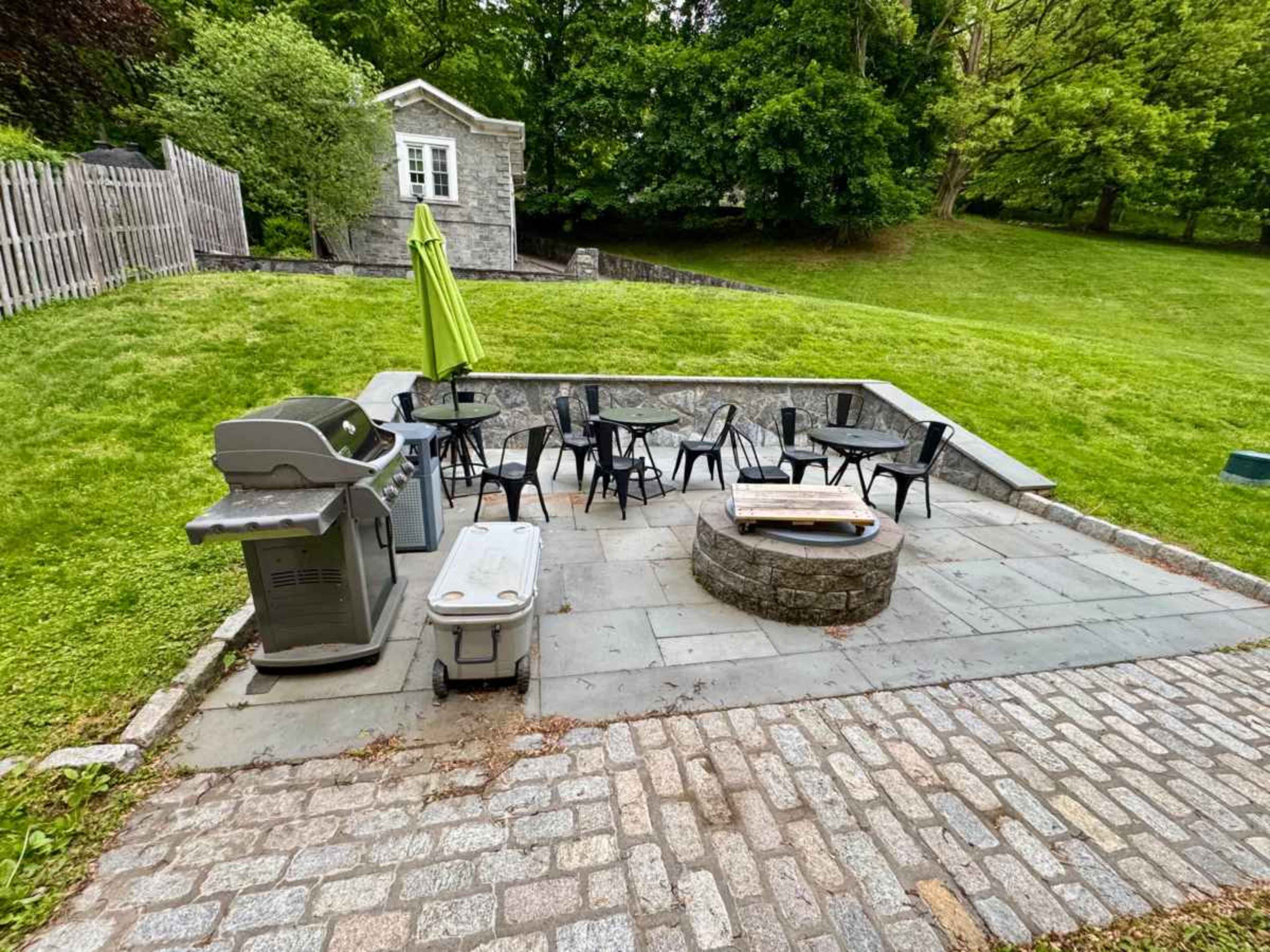 The image shows an outdoor patio area featuring a stone fire pit, a grill, several black chairs around a table, and a green umbrella on a grassy hillside.