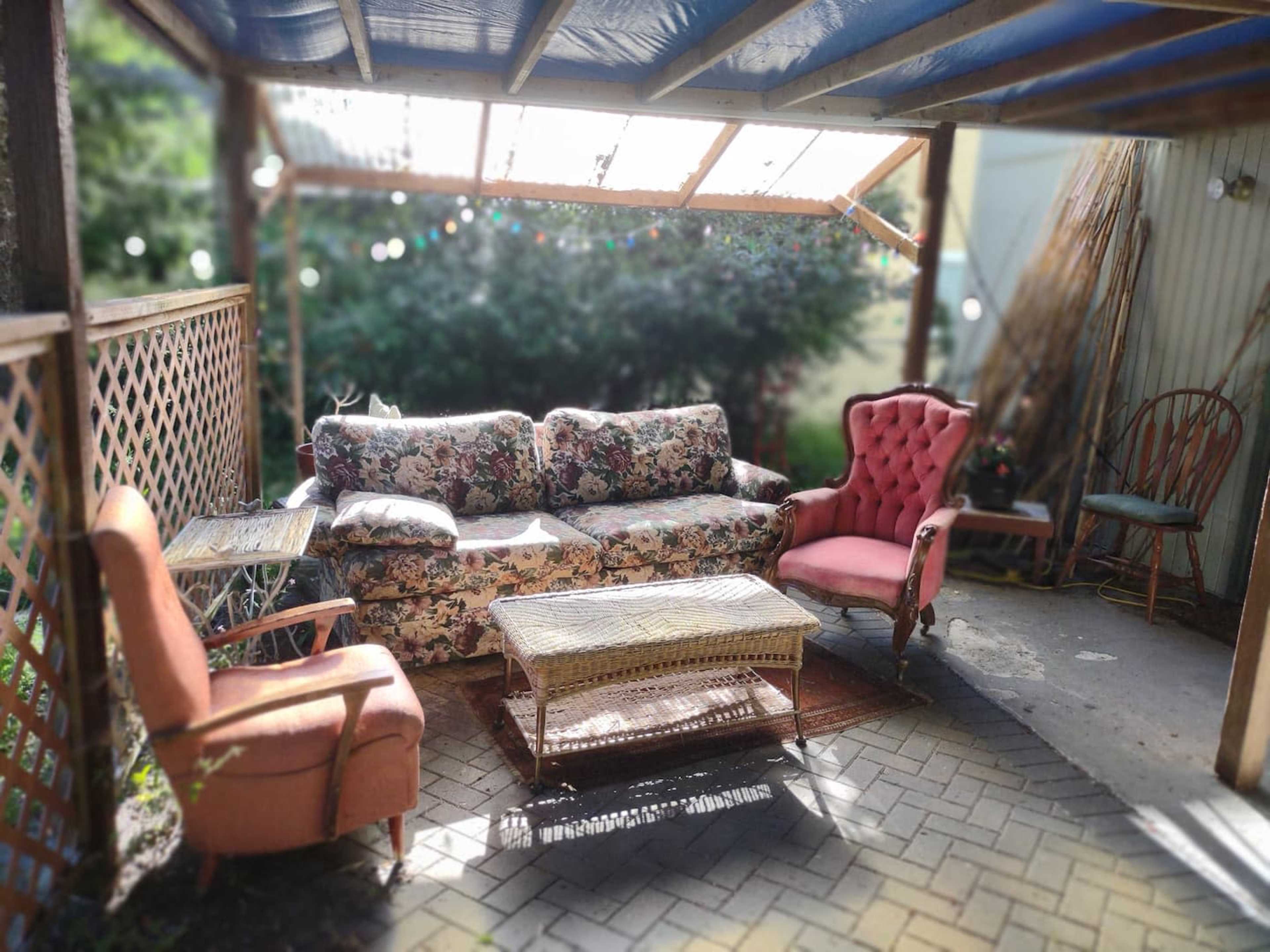 The image shows a cozy outdoor seating area with a floral-patterned couch, two armchairs, and a coffee table under a wooden roof.