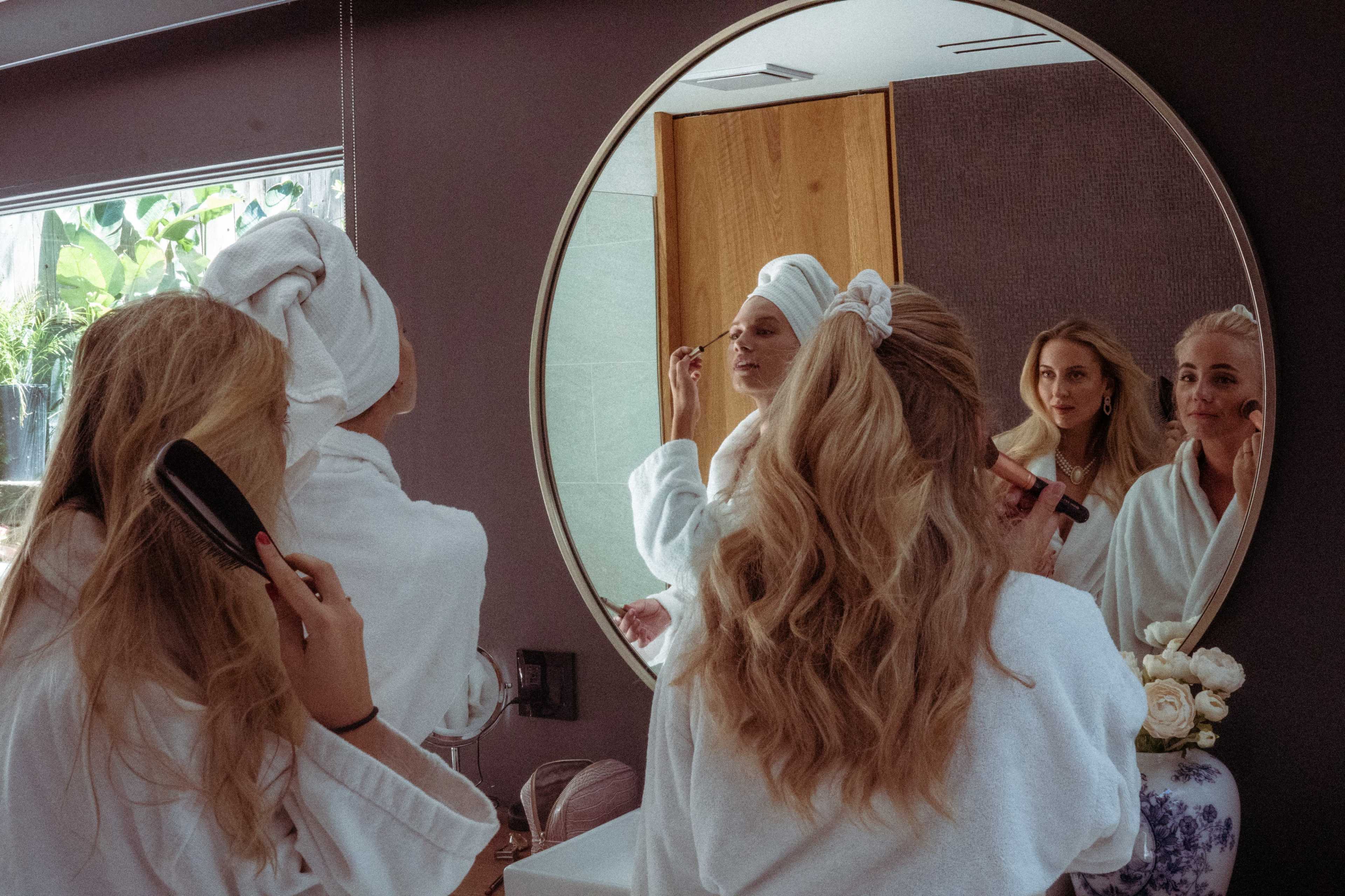 Two women in bathrobes are preparing in front of a mirror, one applying makeup and the other brushing her hair.