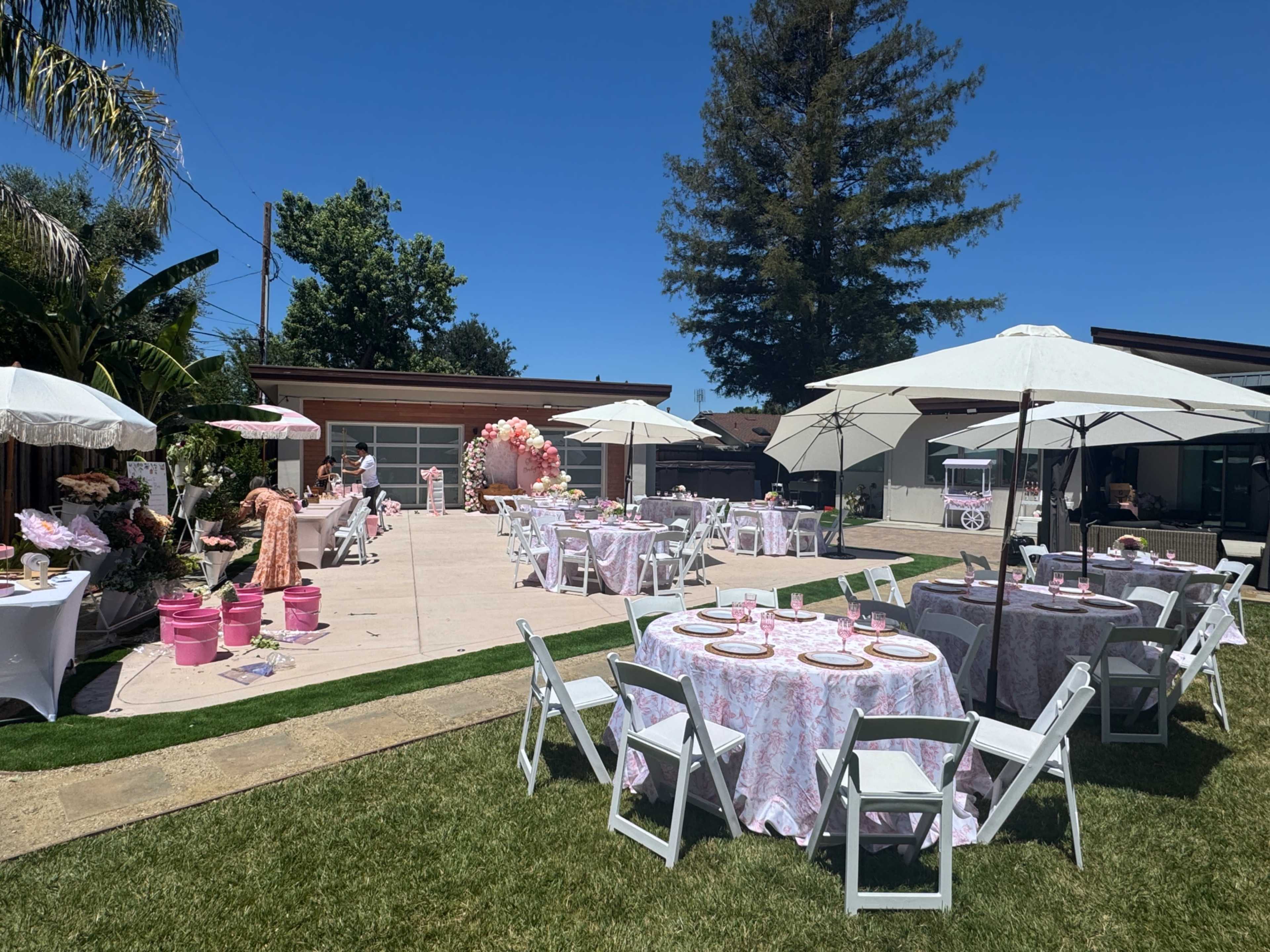 The scene depicts an outdoor event setup with several tables covered in floral tablecloths, white chairs, and large umbrellas, surrounded by greenery and decorations.