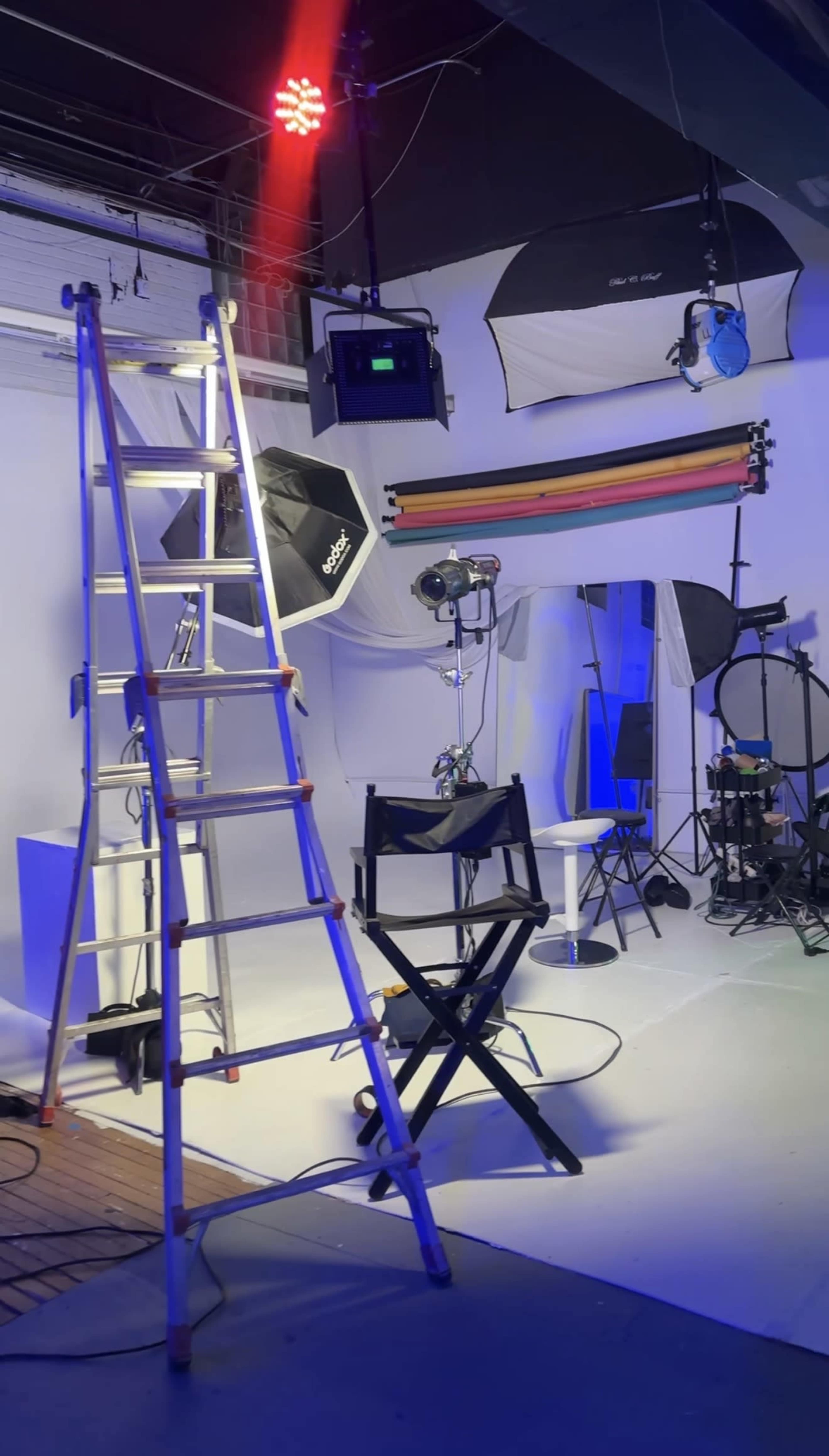 The image shows a photography studio setup with a ladder, a director's chair, various lighting equipment, and backdrops arranged in a brightly lit environment.