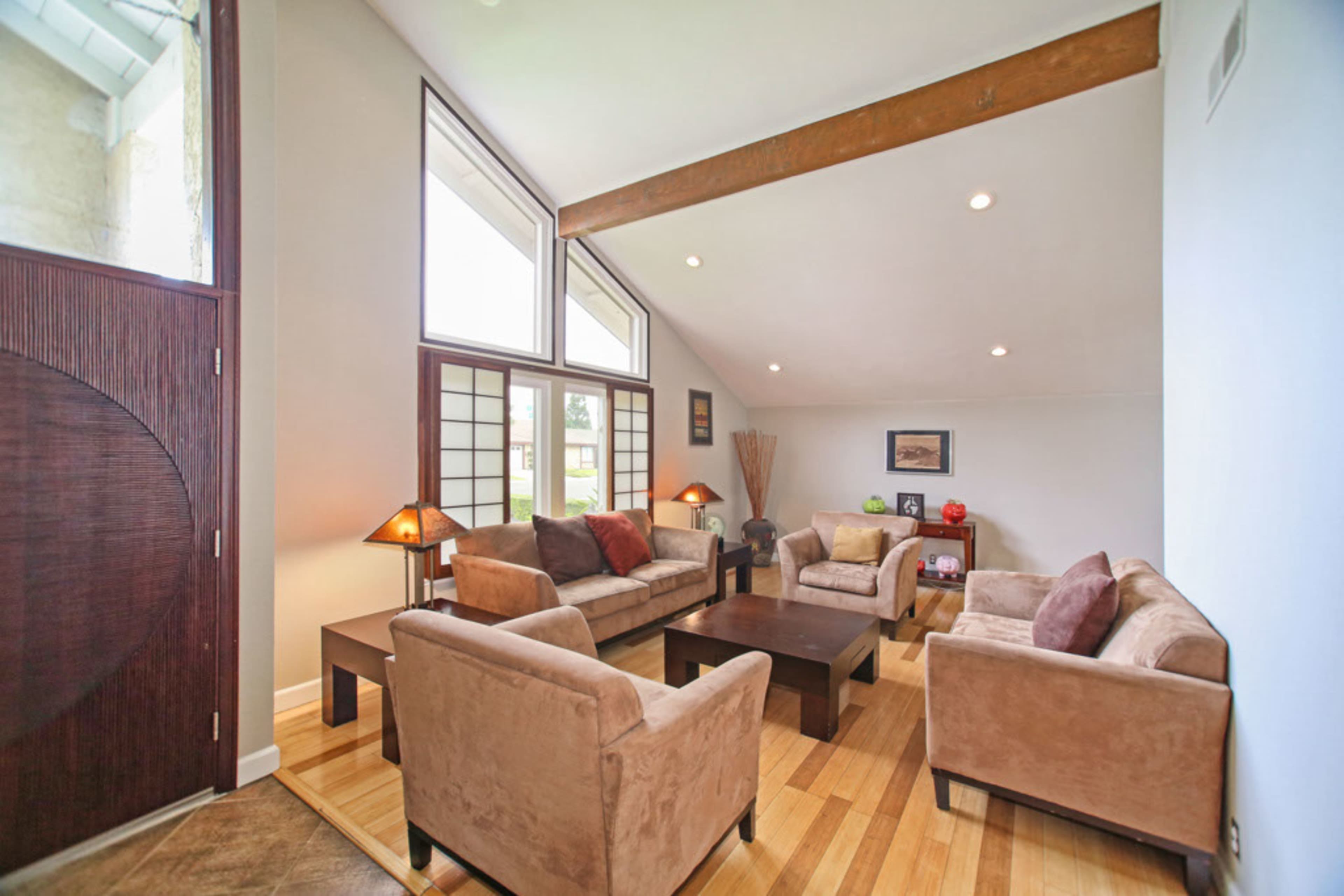 The image shows a bright living room with large windows, featuring a seating arrangement of beige sofas and chairs around a dark coffee table.