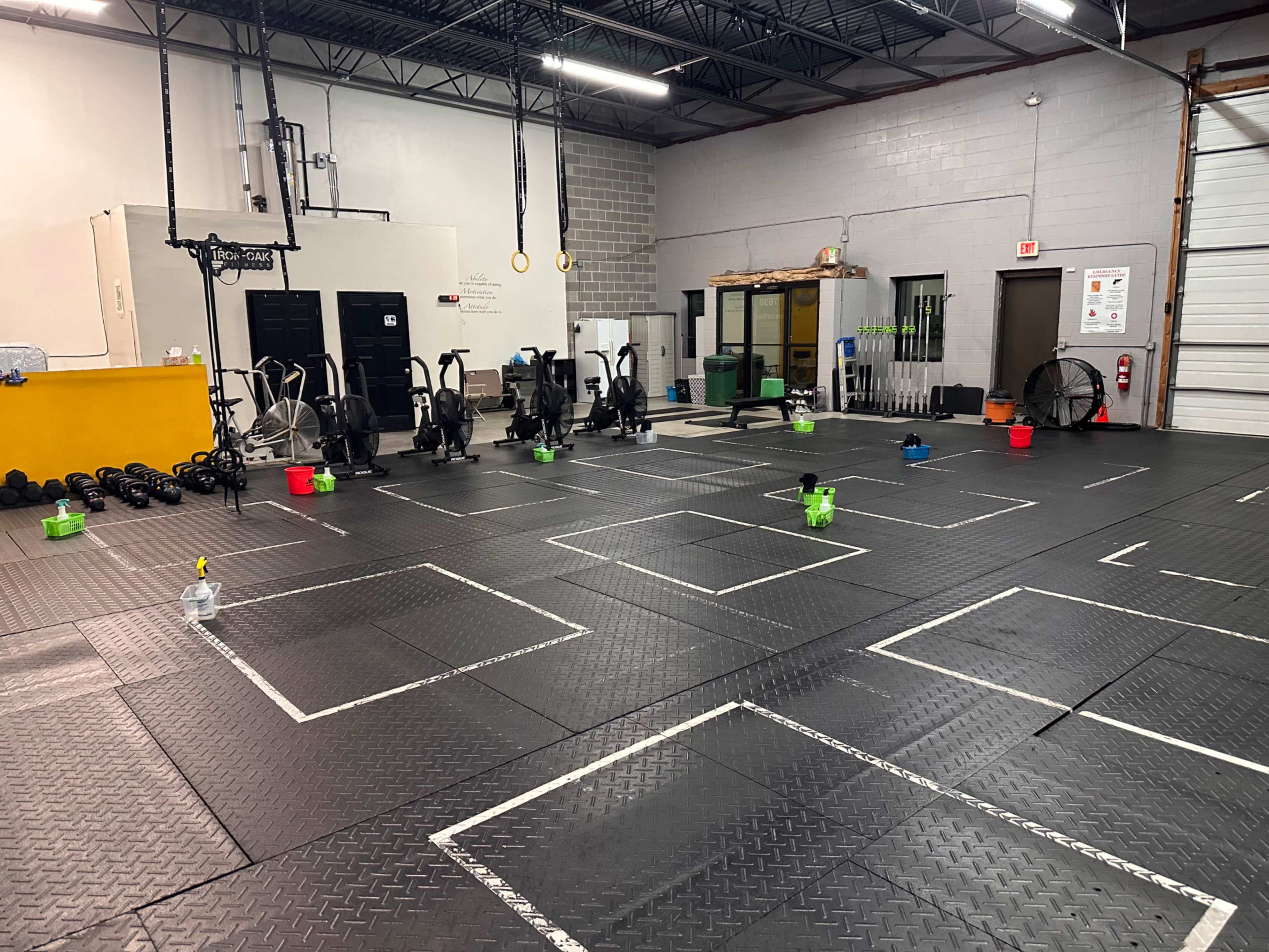A spacious gym with exercise equipment, including stationary bikes and weights, arranged in sections defined by white tape on a black rubber floor.