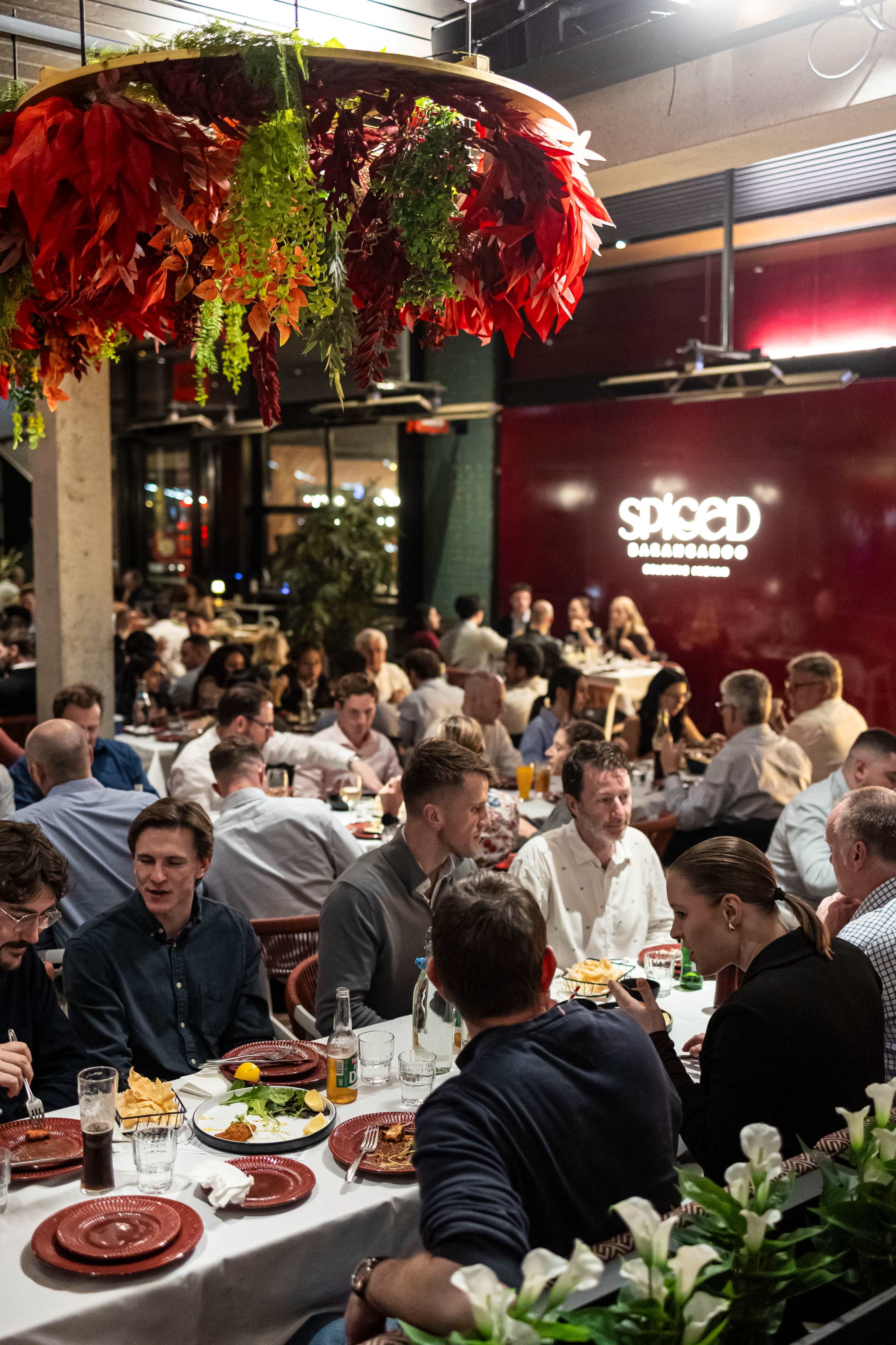 A crowded restaurant features people dining at tables adorned with food and drinks under vibrant hanging decorations.