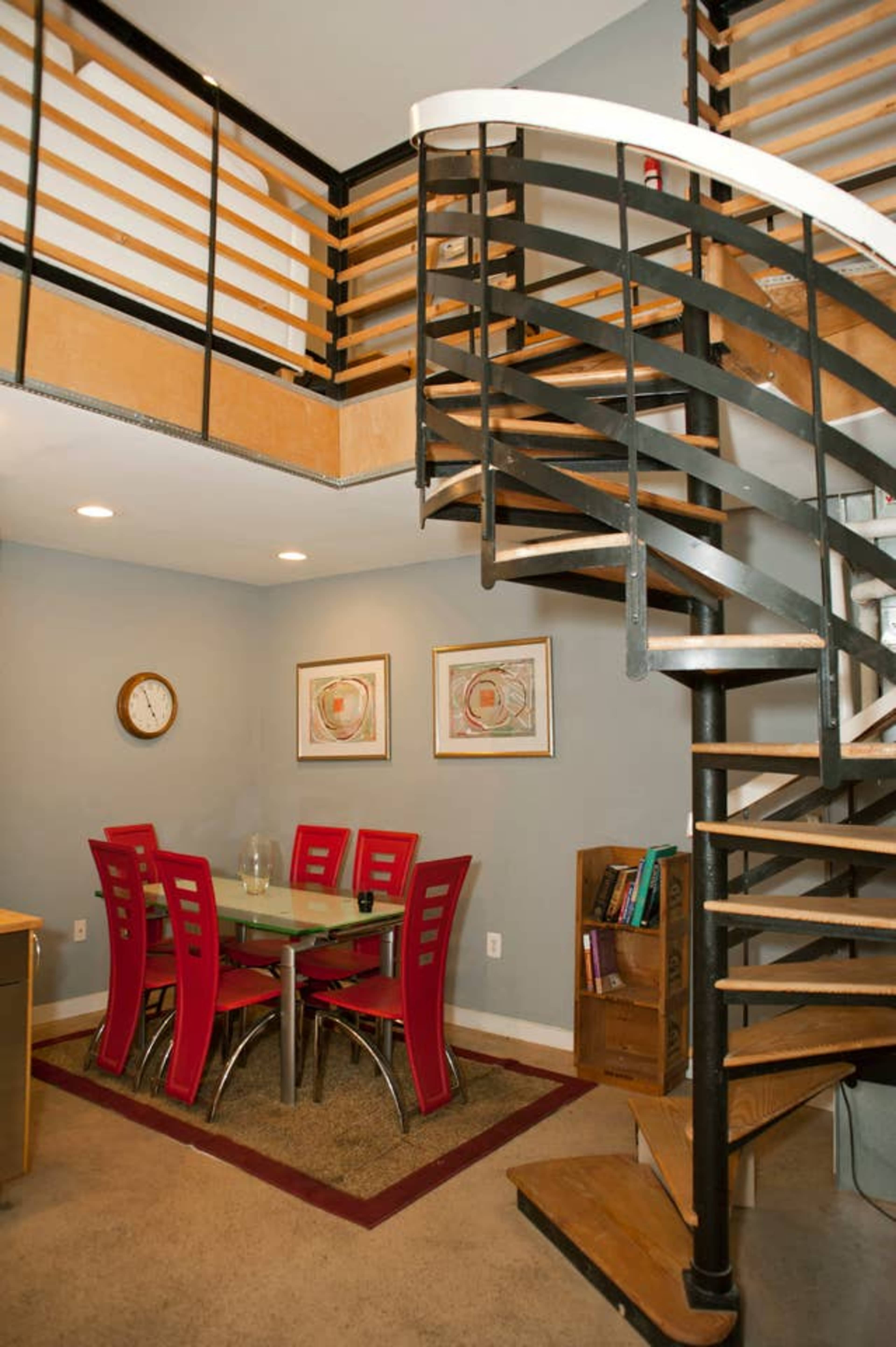 The image depicts a spiral staircase leading to an upper level, with a dining area featuring a glass table and red chairs on a gray floor.