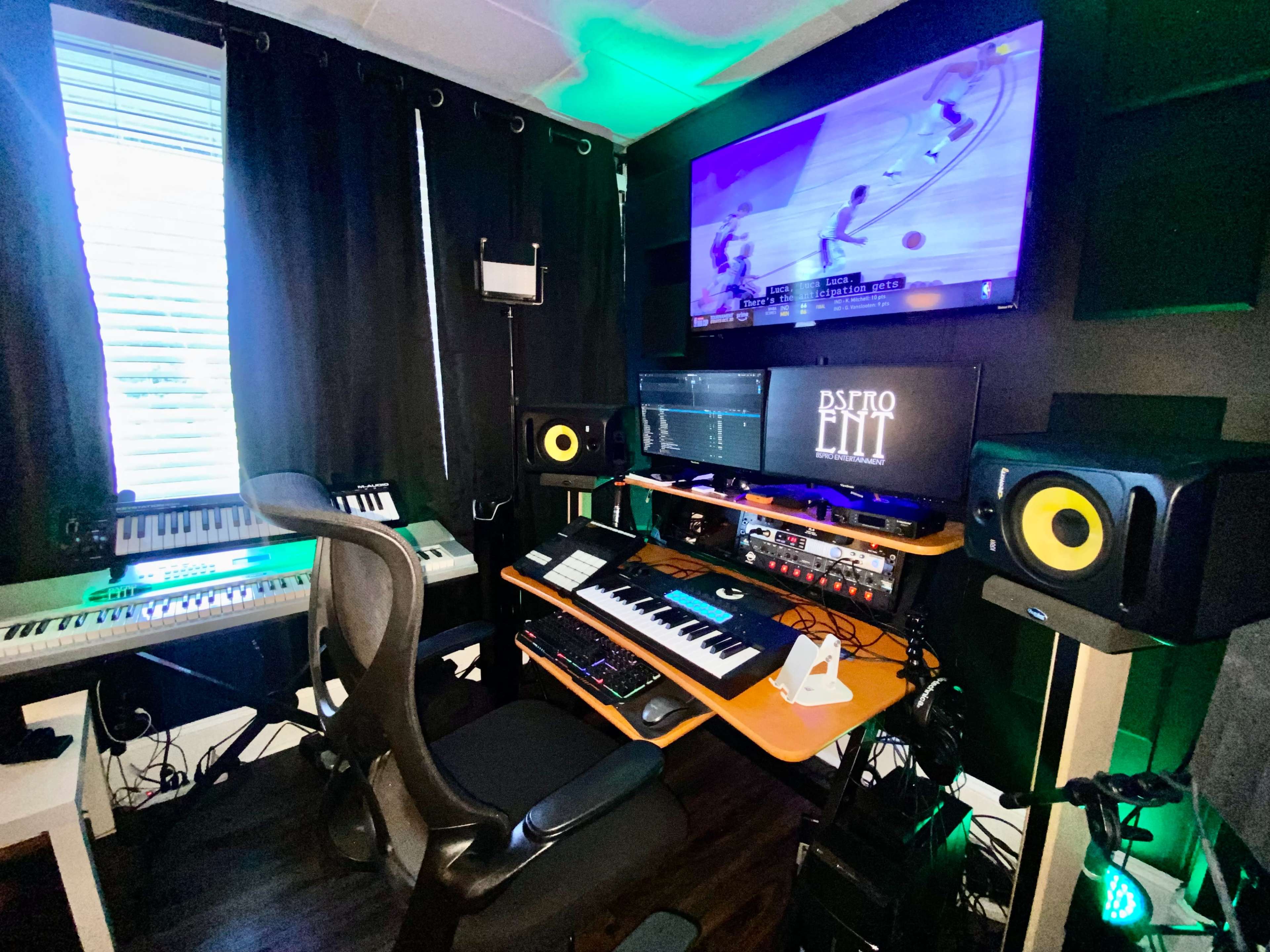 The image shows a music production studio featuring multiple keyboards, a computer setup with monitors, and a wall-mounted TV displaying a basketball game.