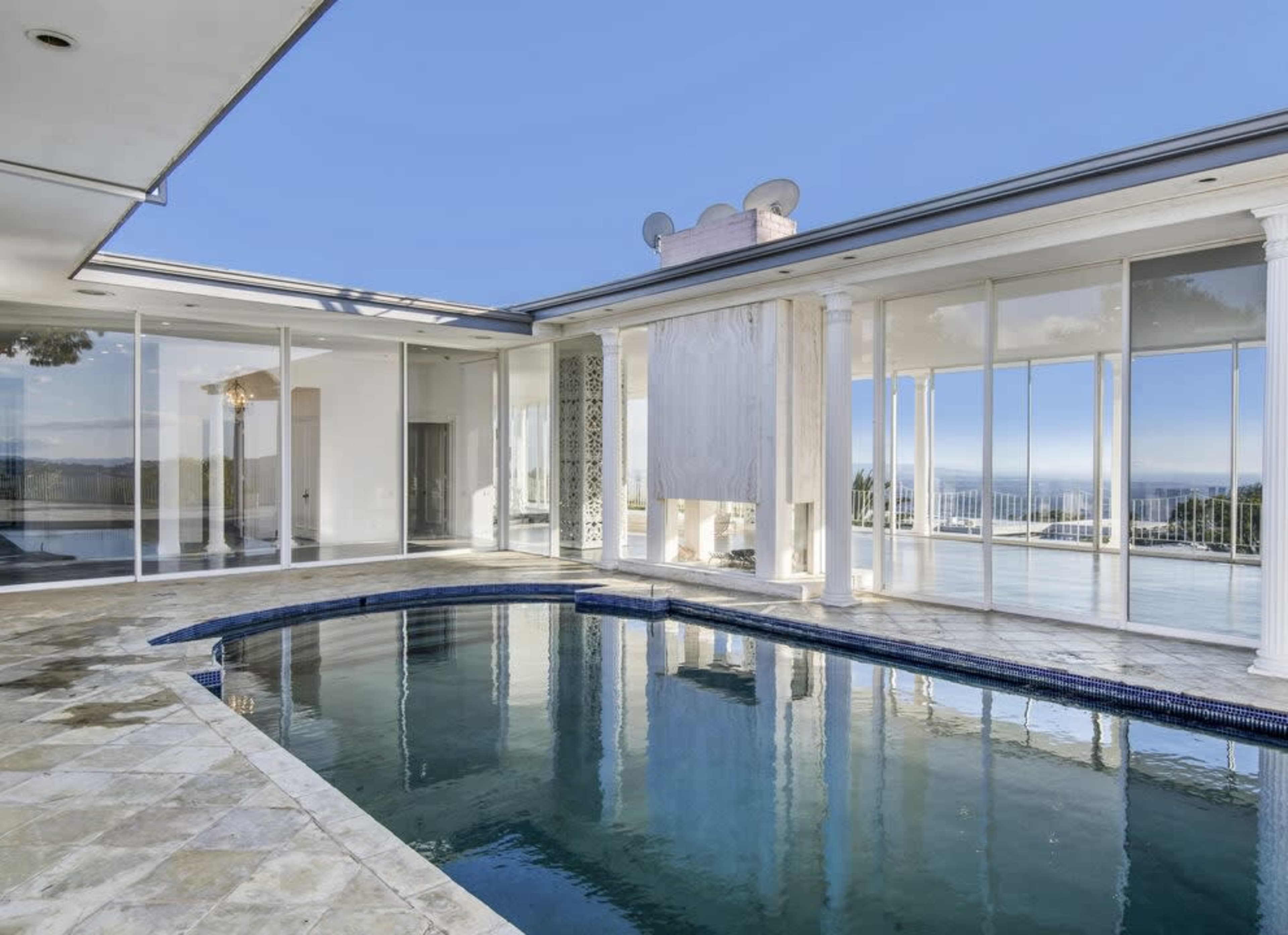 The image shows a modern interior with a pool, large glass windows, and columns, overlooking a scenic view.