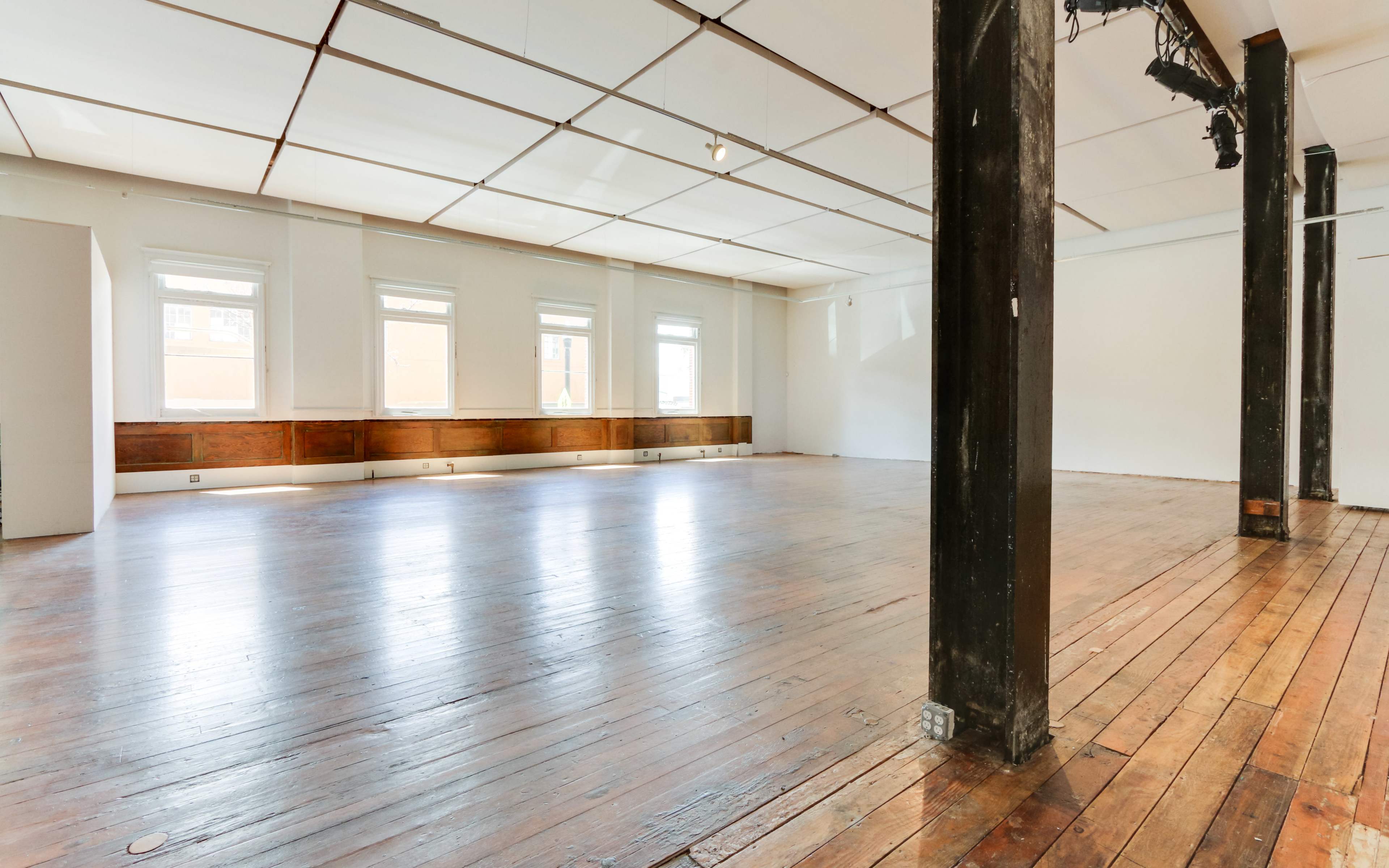 A spacious, empty room with wooden flooring, large windows, and exposed beams.