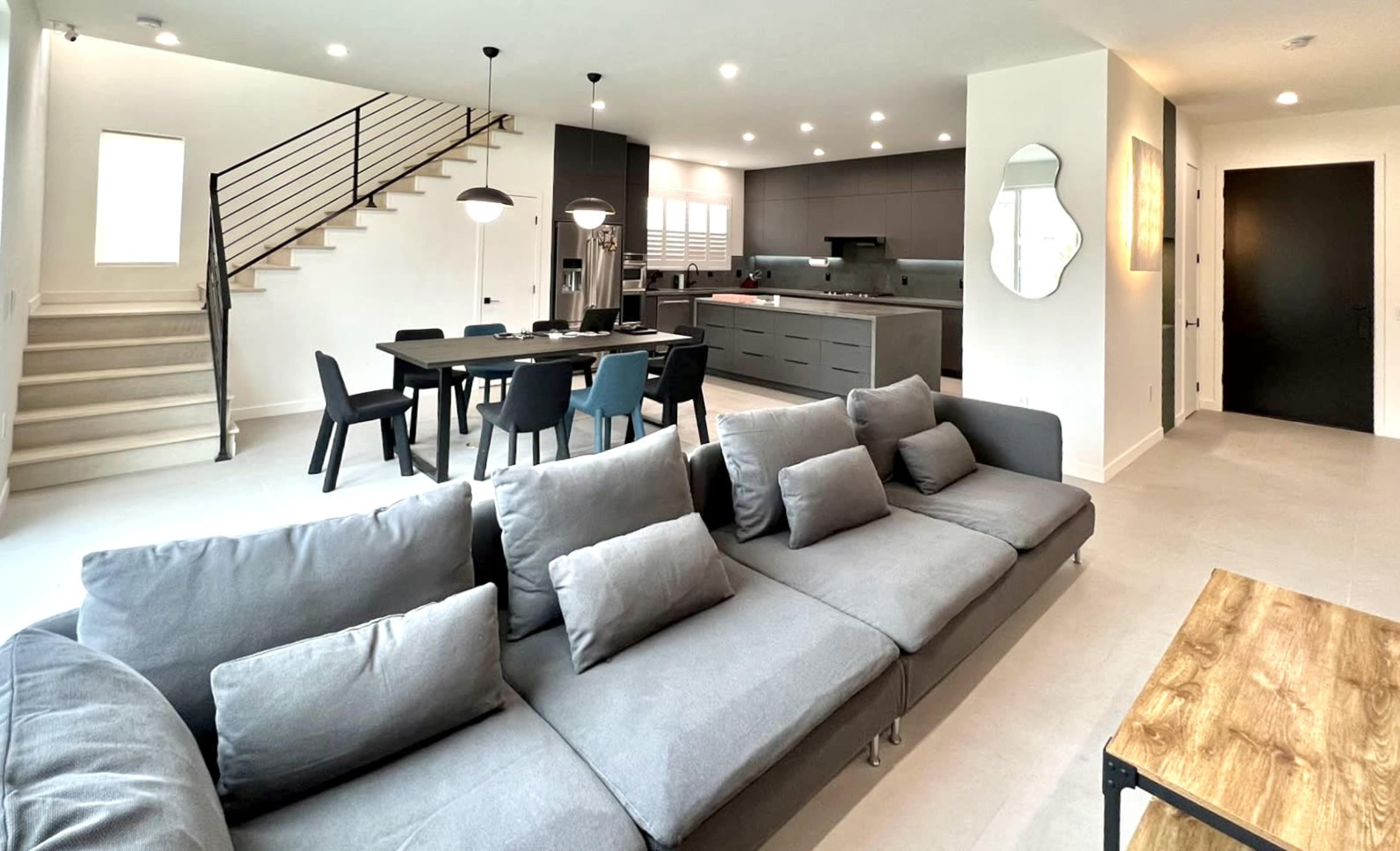 A modern living space with a grey sofa in the foreground, a dining area with a black table and chairs, and a kitchen featuring sleek cabinetry and appliances.
