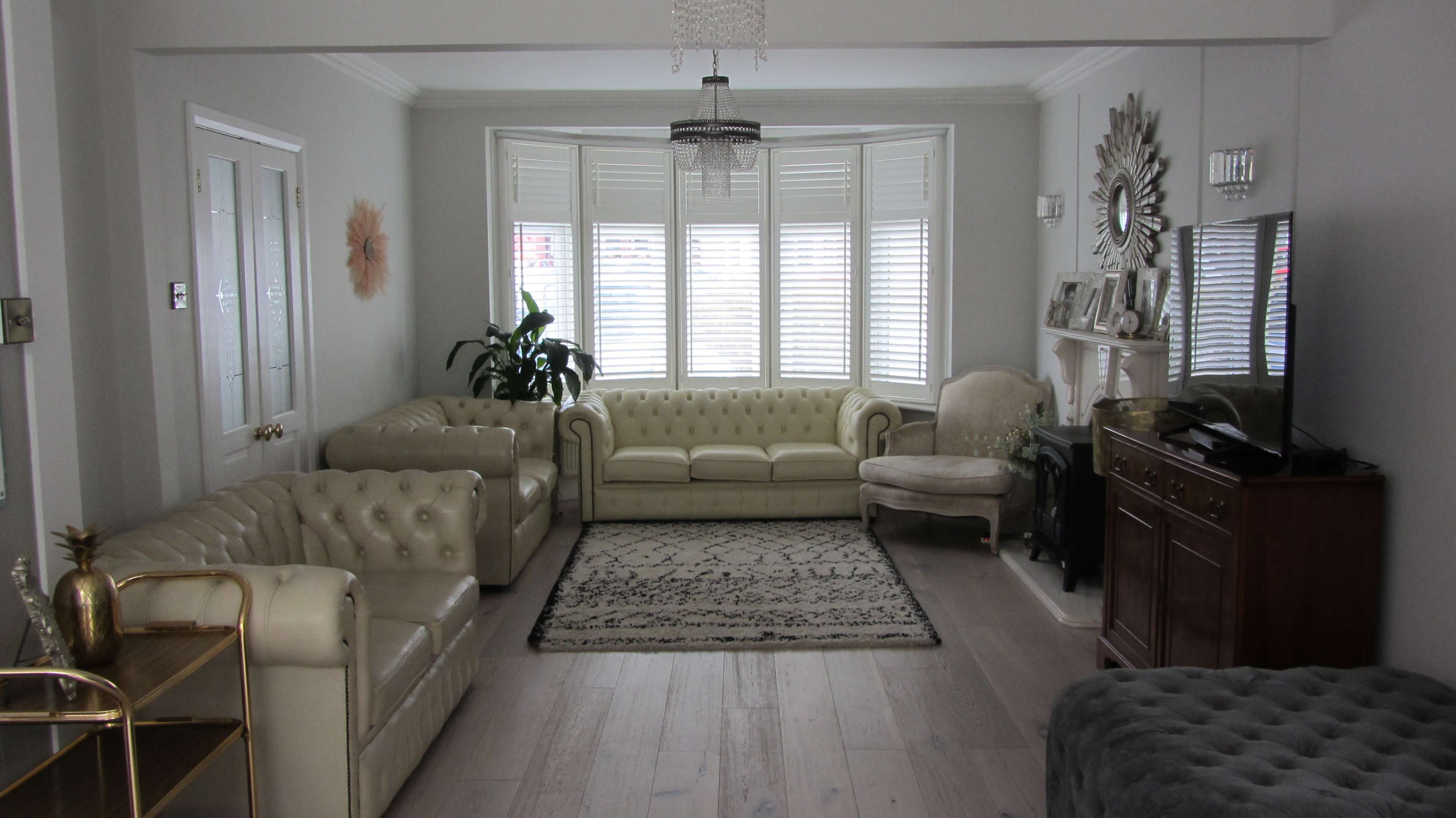 A well-furnished living room features two cream-colored tufted sofas, a large area rug, and a bay window with shuttered blinds.
