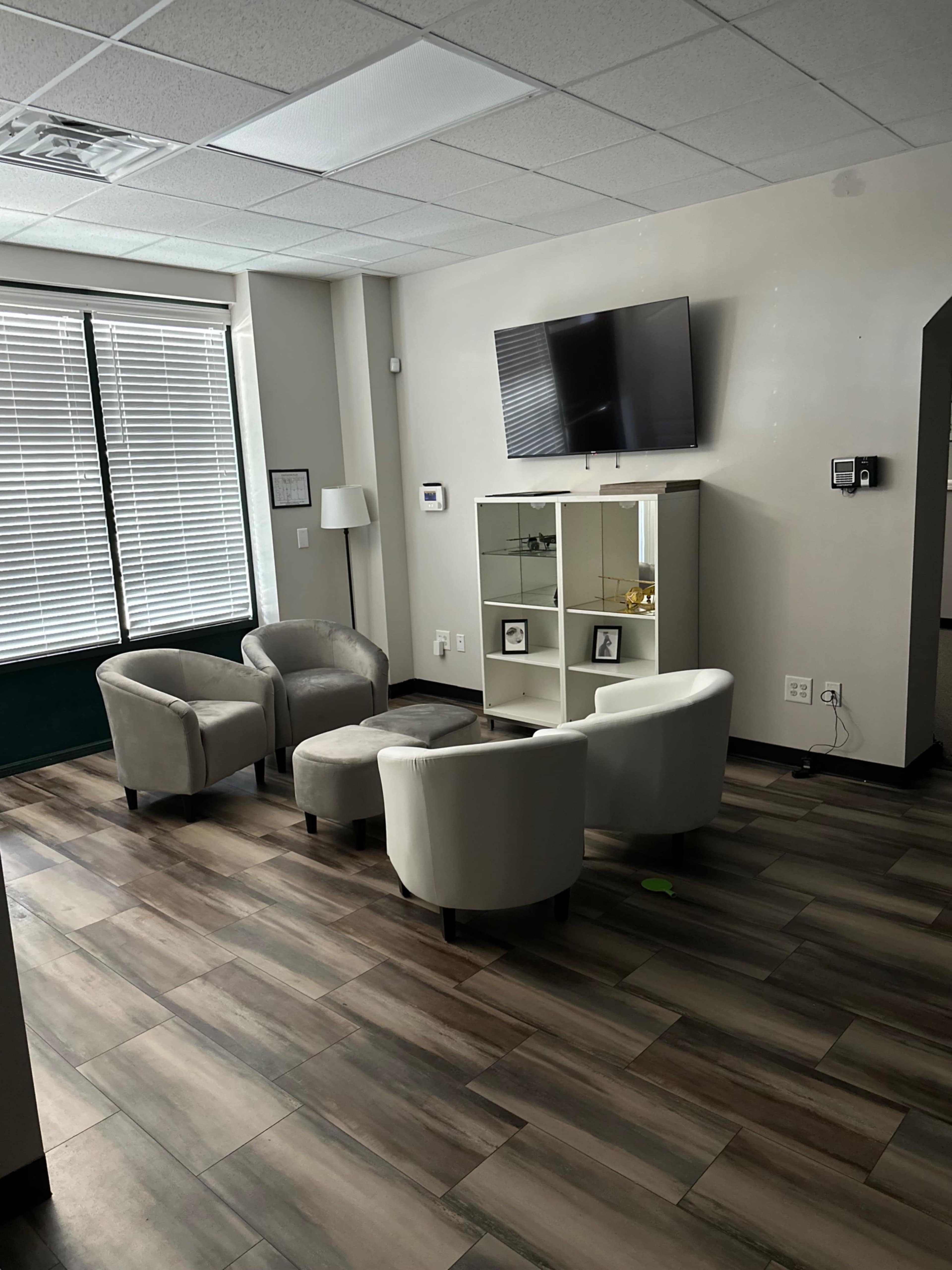 A waiting area with four light-colored chairs arranged around a small ottoman, a television mounted on the wall, and a glass cabinet displaying picture frames.