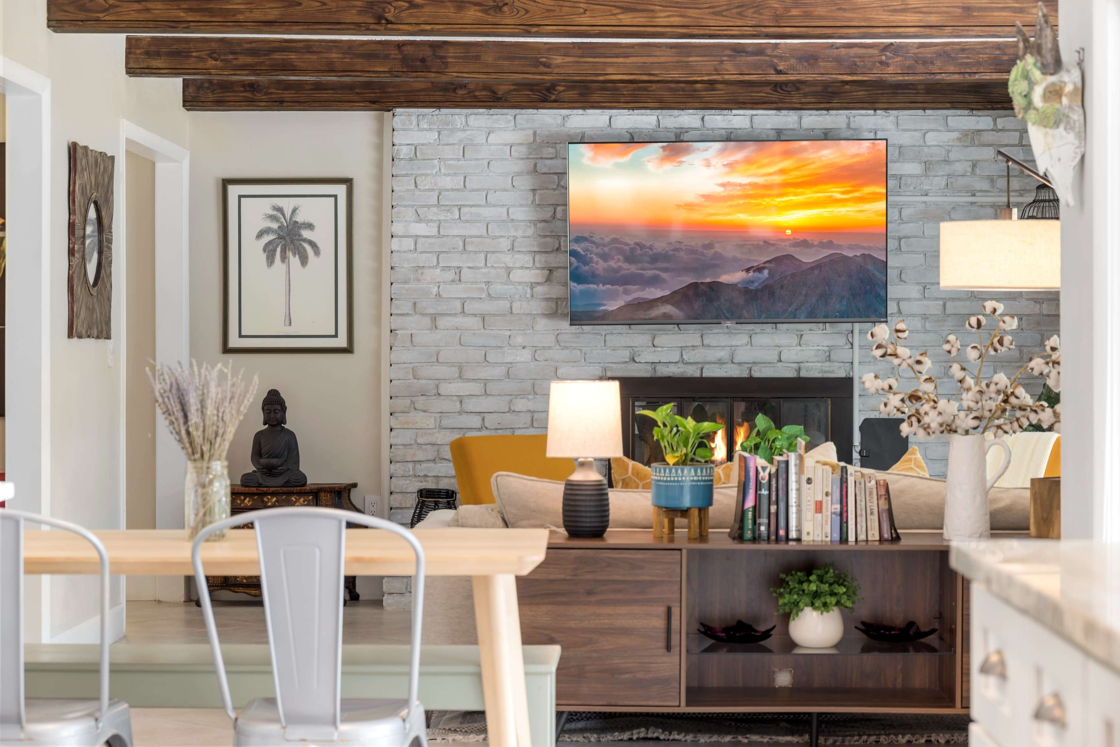 A cozy living room features a mounted television displaying a sunset over mountains, with a yellow couch, a coffee table, and a decorative bookshelf.