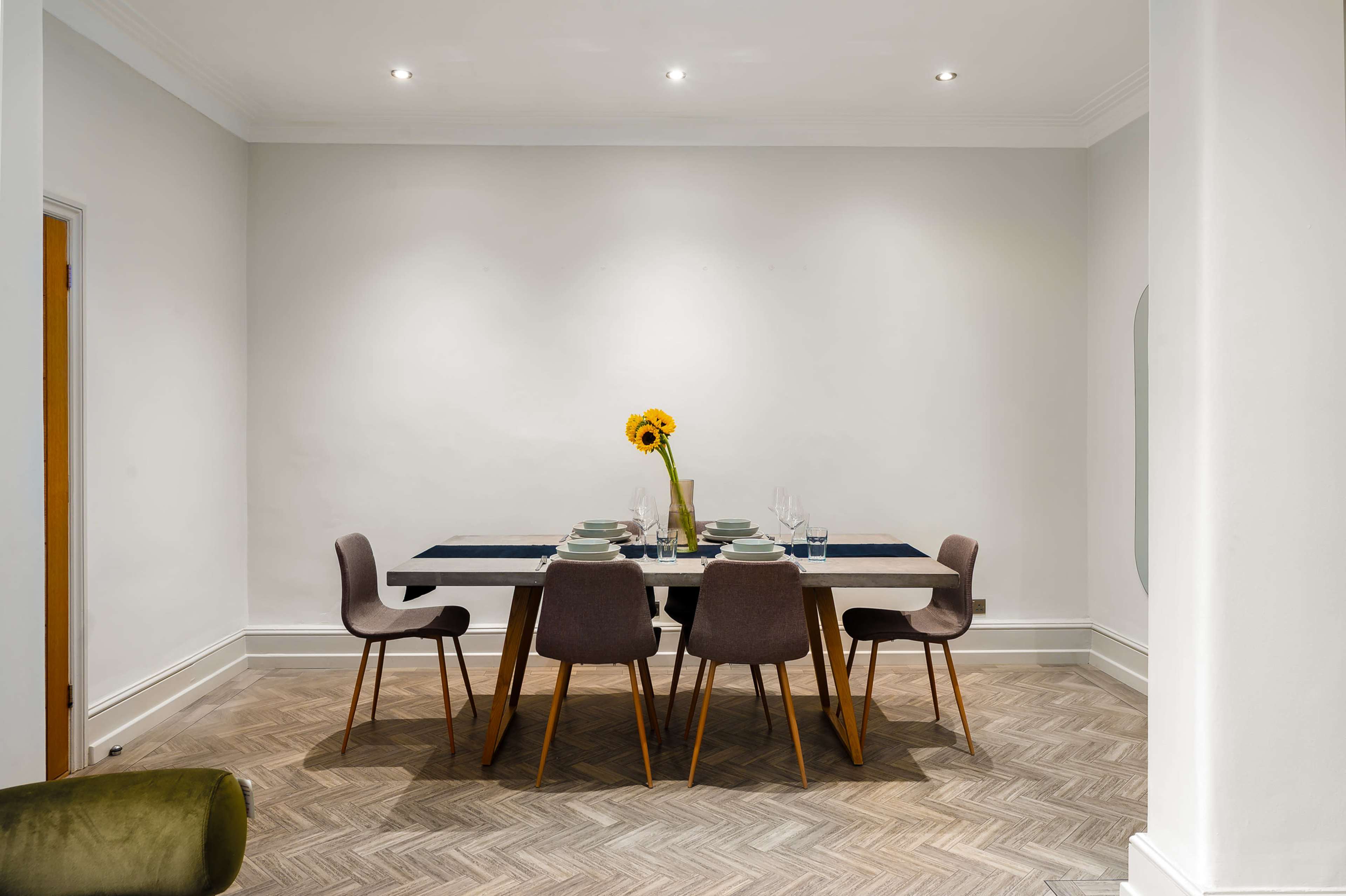 A dining table set for a meal with plates and glasses, accompanied by a sunflower vase, is surrounded by four chairs in a modern room with hardwood flooring.