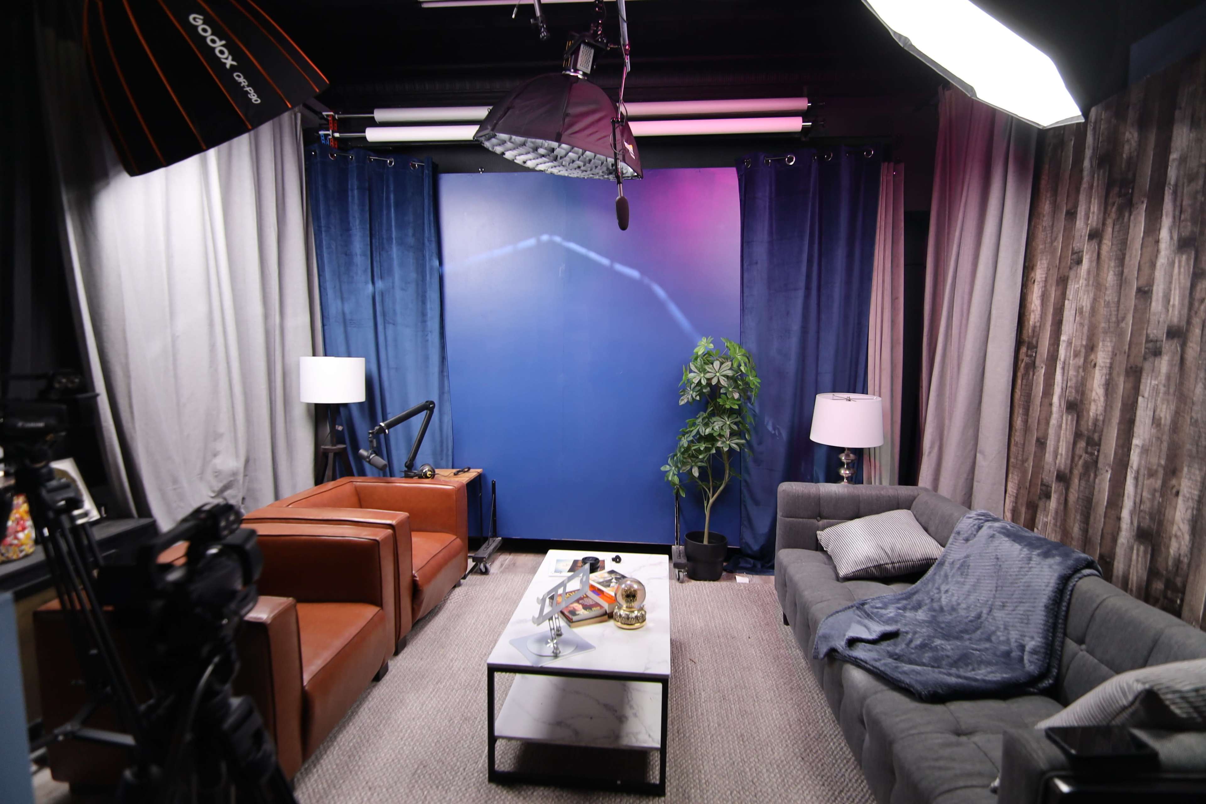 A well-lit studio setup with two armchairs, a sofa, a coffee table, and a backdrop of blue curtains and lighting equipment.