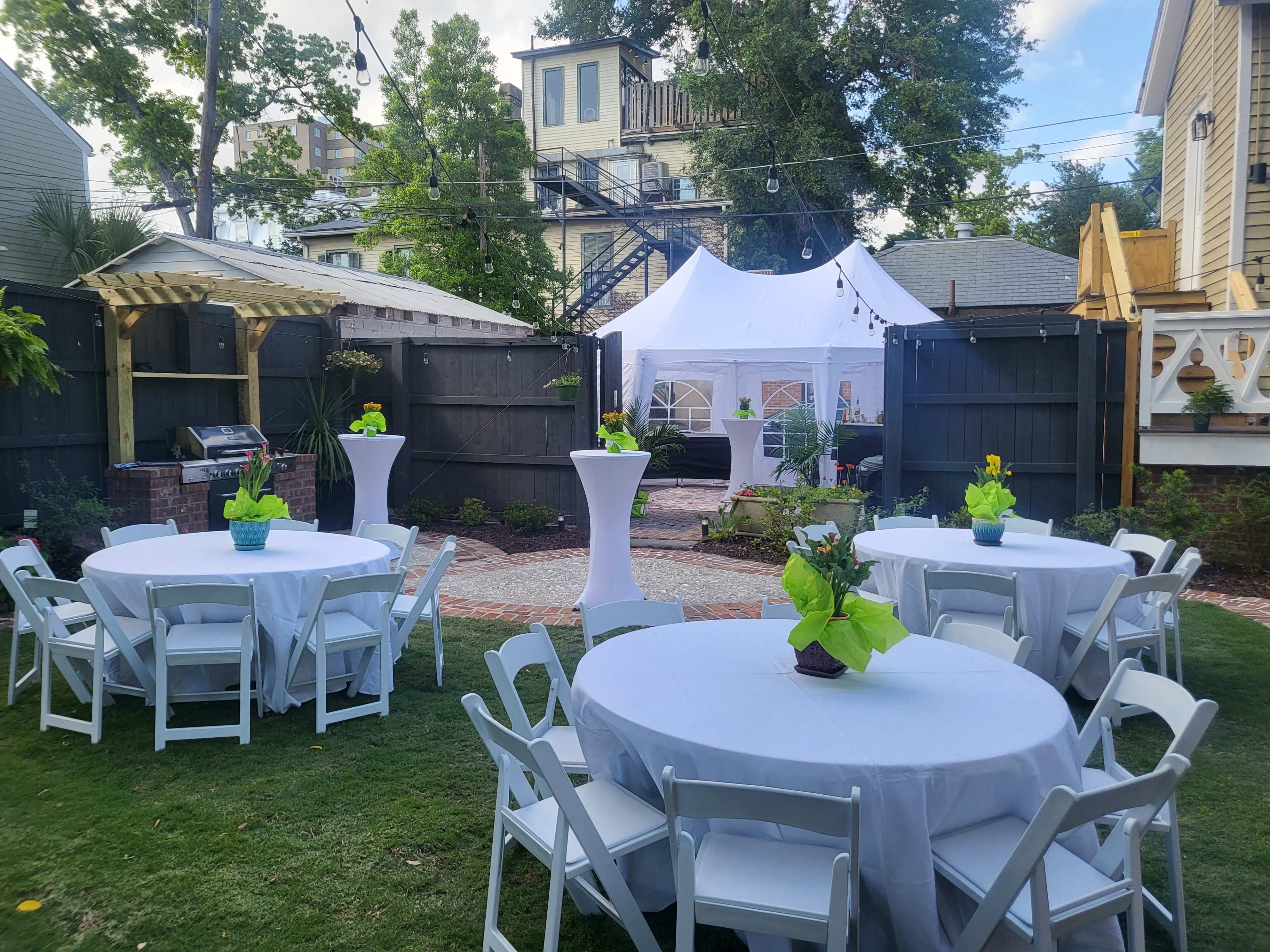 A backyard setup for an event, featuring several round tables with white tablecloths, each adorned with potted plants, and a white tent in the background.