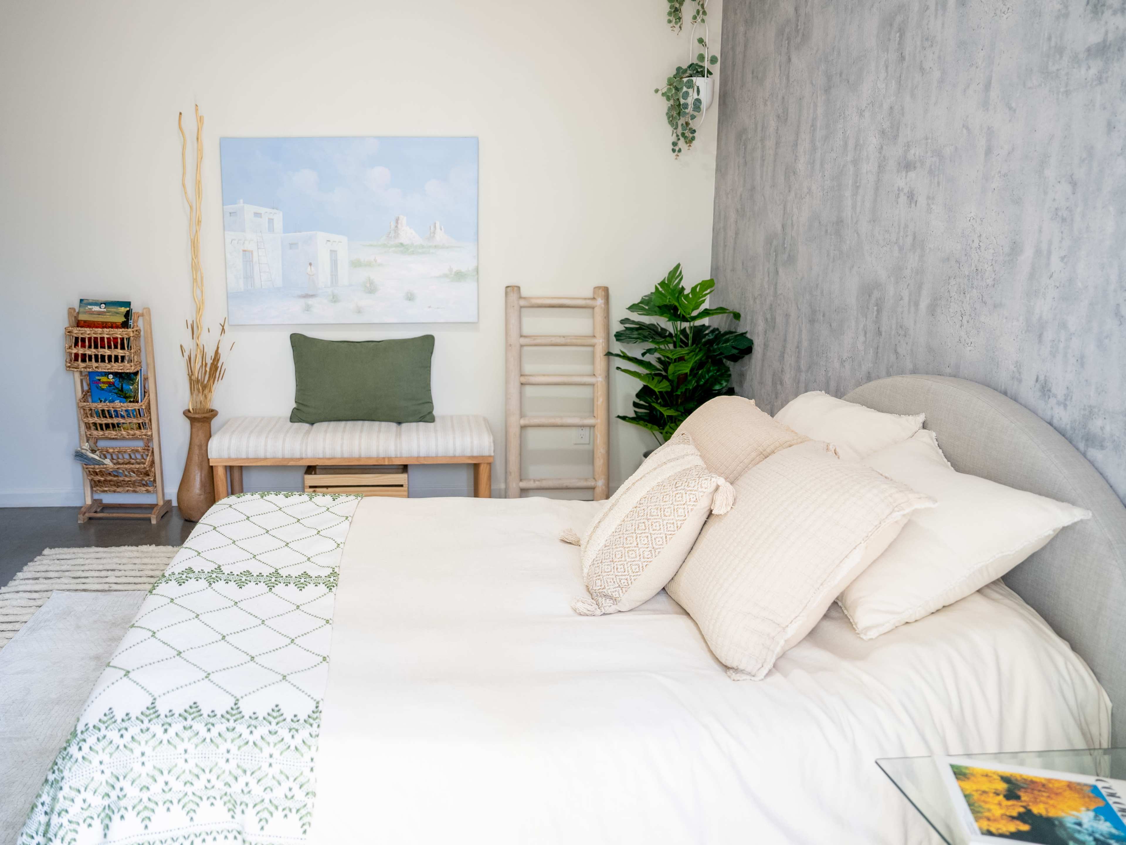 A well-organized bedroom features a large bed with light linens, a wooden ladder against the wall, and a painting above a bench.