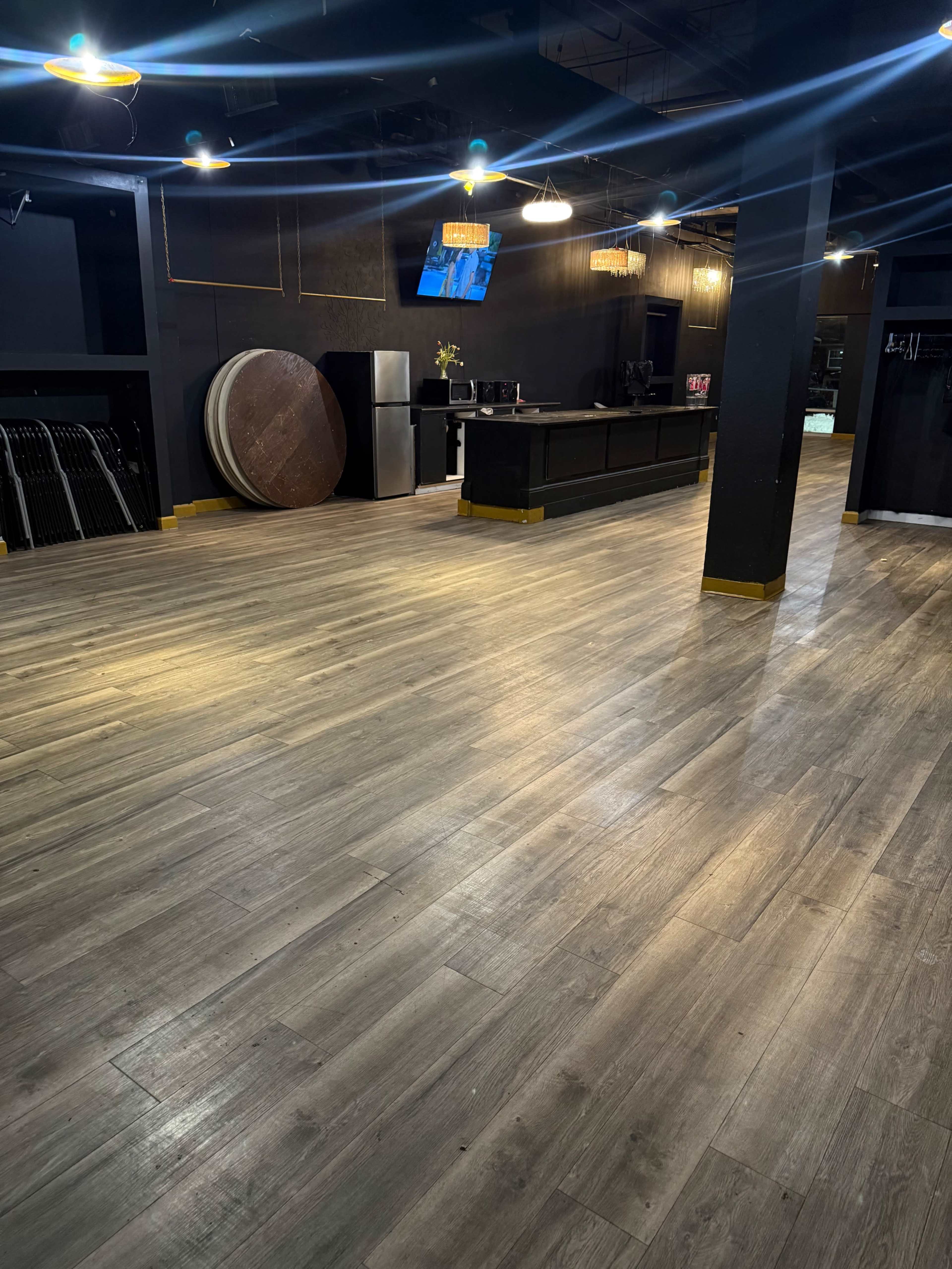 The image shows an empty interior space with wooden flooring, a bar area in the back, and various objects like tables and a refrigerator visible in the background.