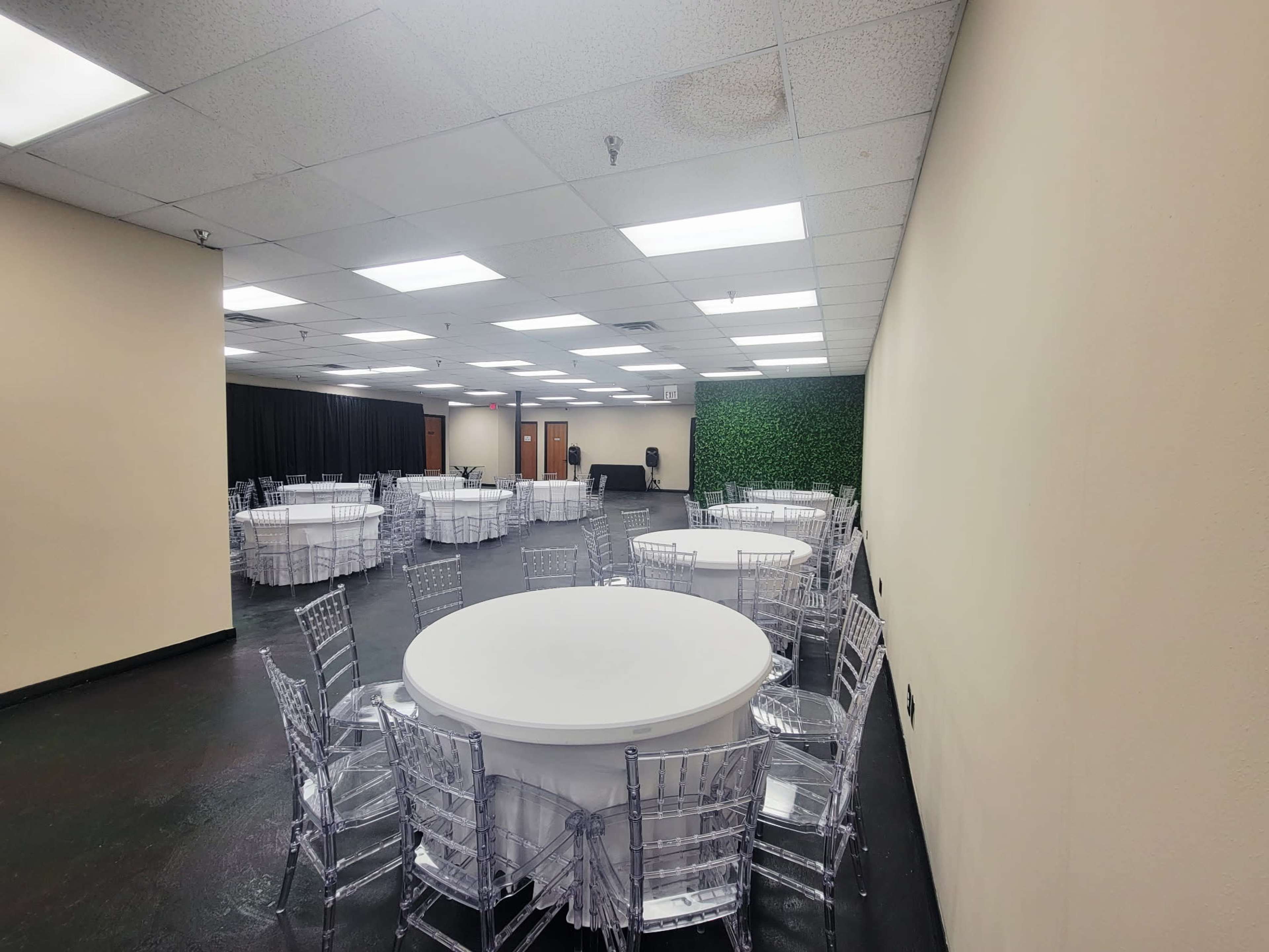 The image shows a spacious event room with several round tables surrounded by transparent chairs, illuminated by overhead lighting.