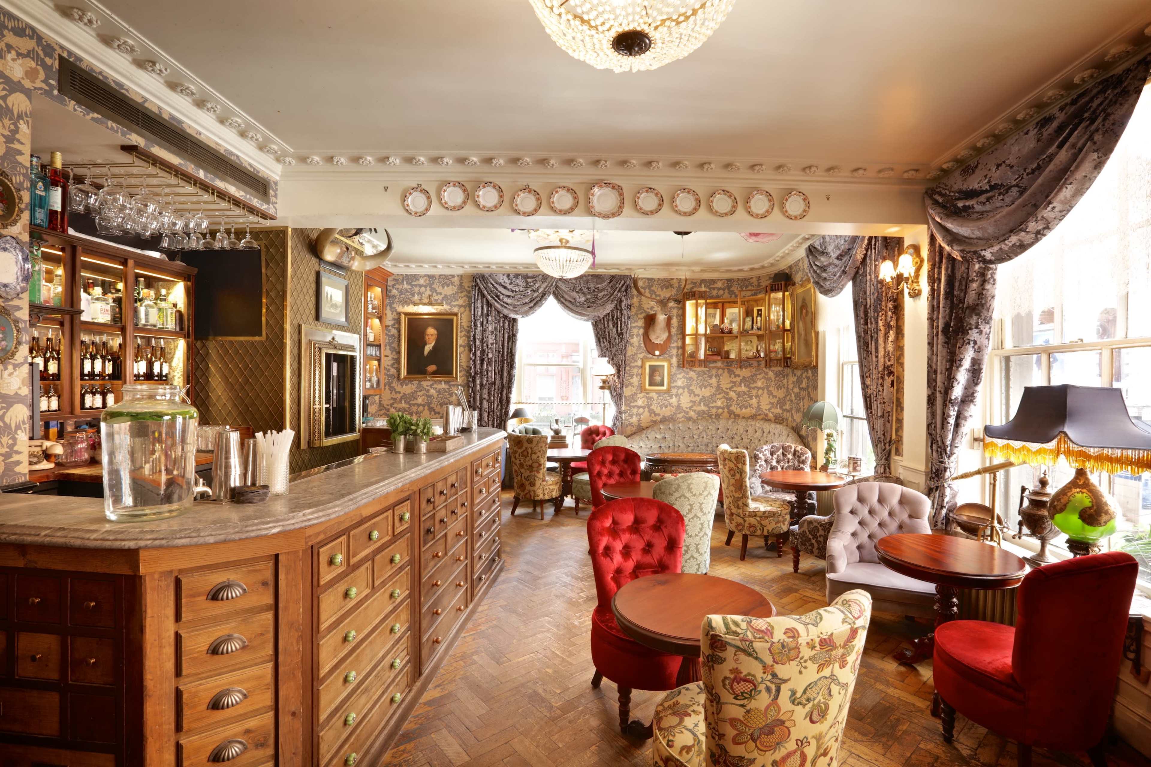 A cozy pub interior features a wooden bar, ornate wallpaper, and various seating areas with patterned chairs.