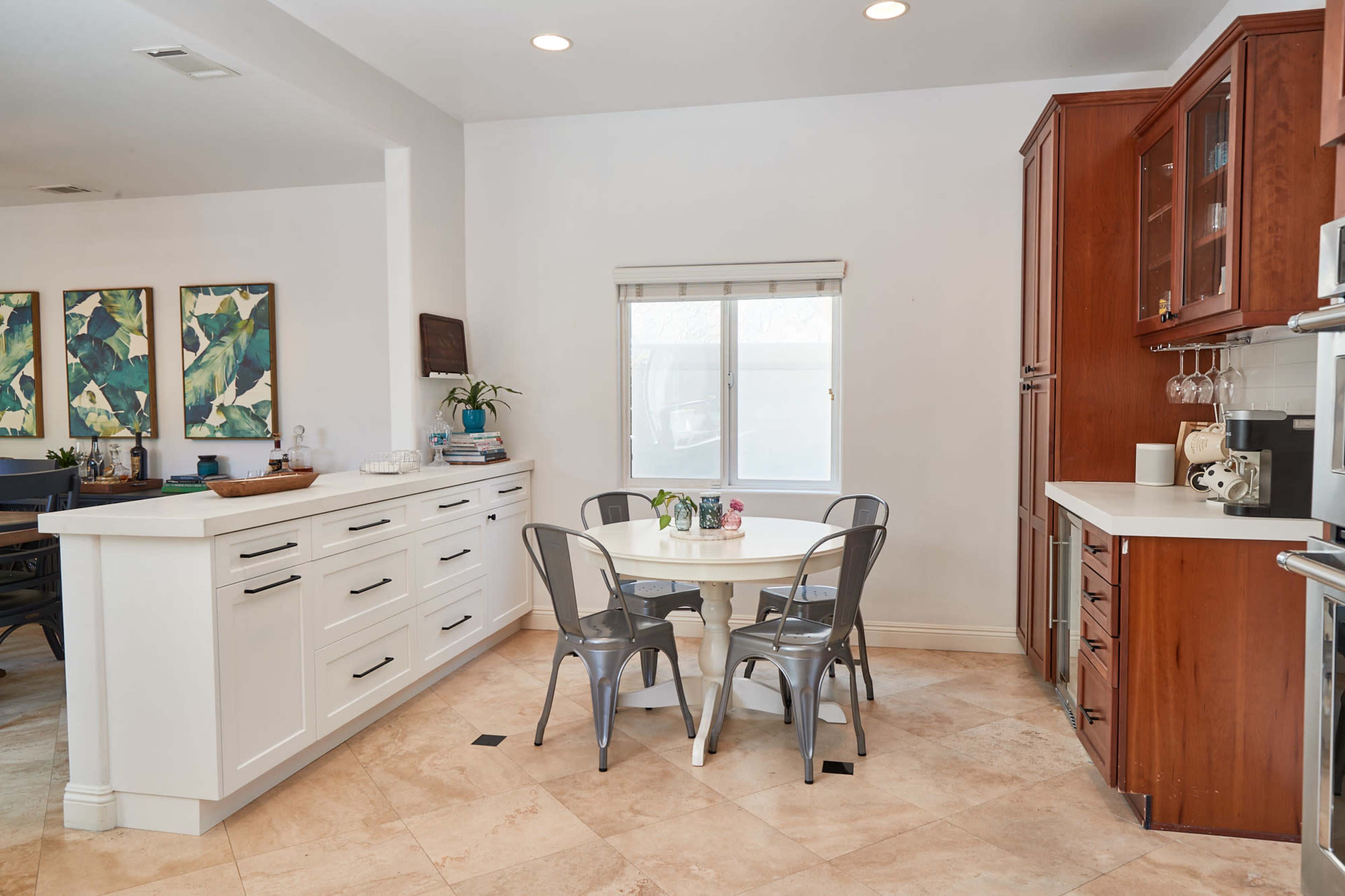 A bright kitchen features white cabinets, a round table with metal chairs, and large windows allowing natural light.