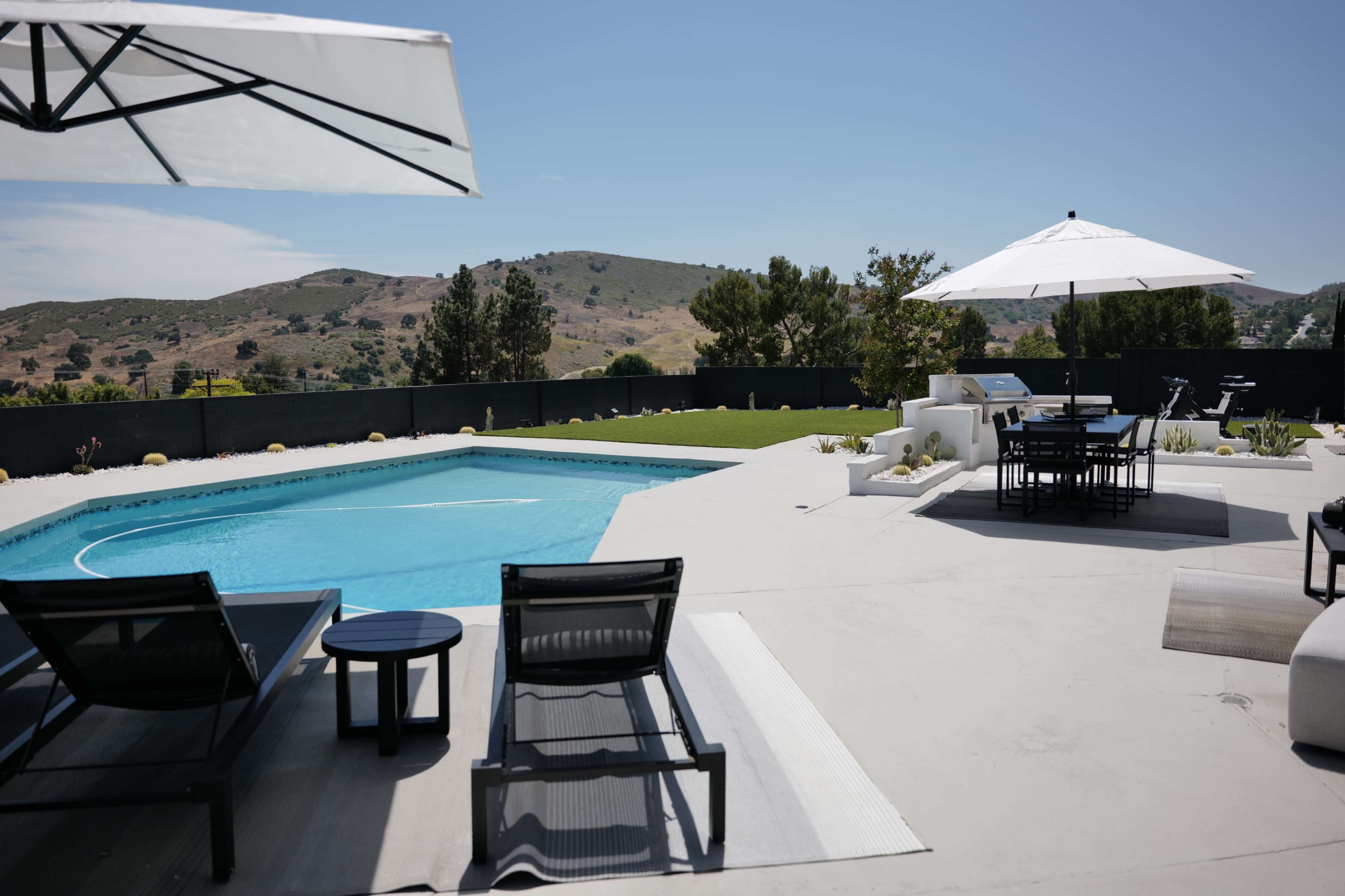 A swimming pool is surrounded by patio chairs and umbrellas, with a view of green hills in the background.