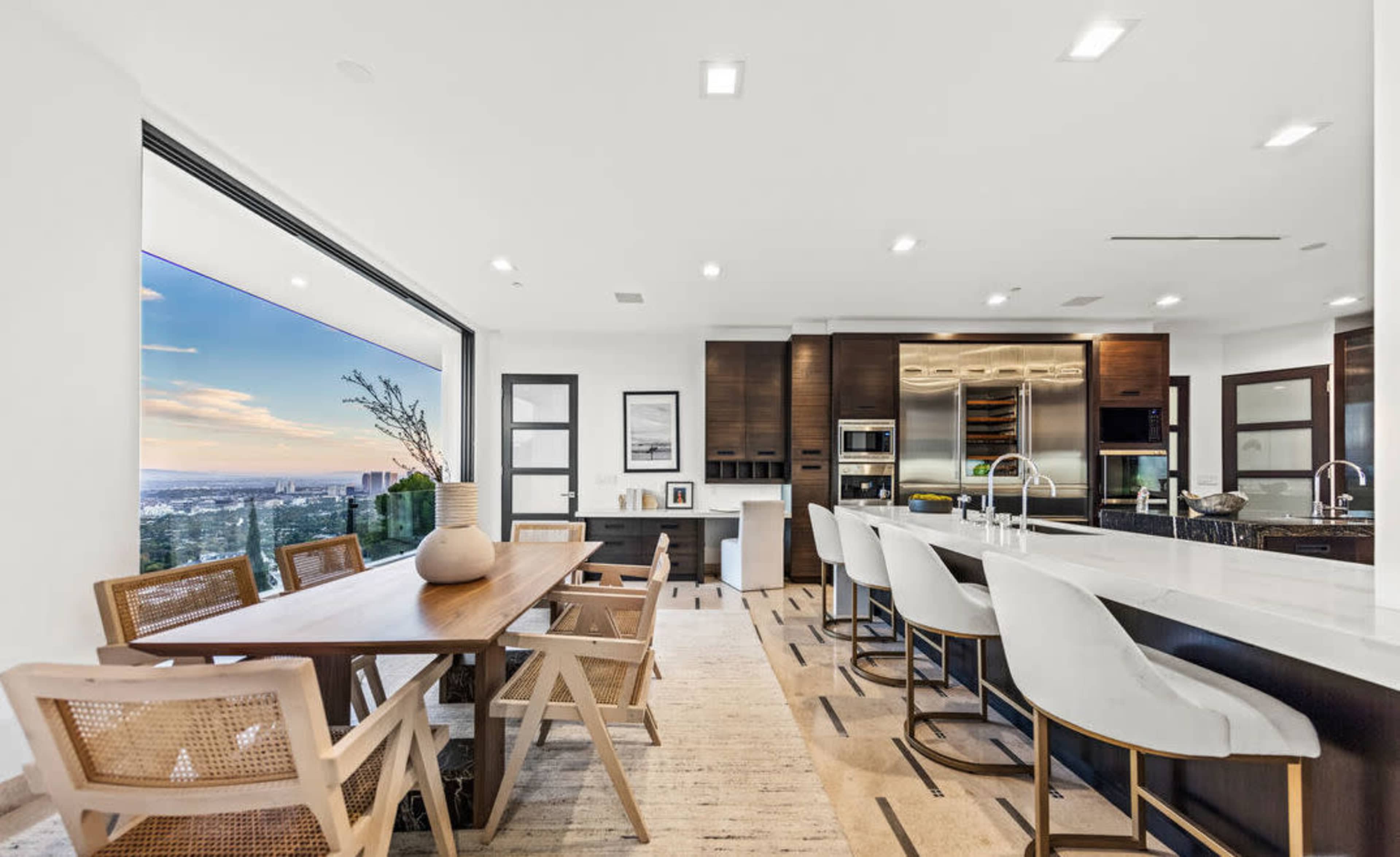 The image shows a modern kitchen and dining area featuring a large table, bar seating, and a panoramic window with a view.