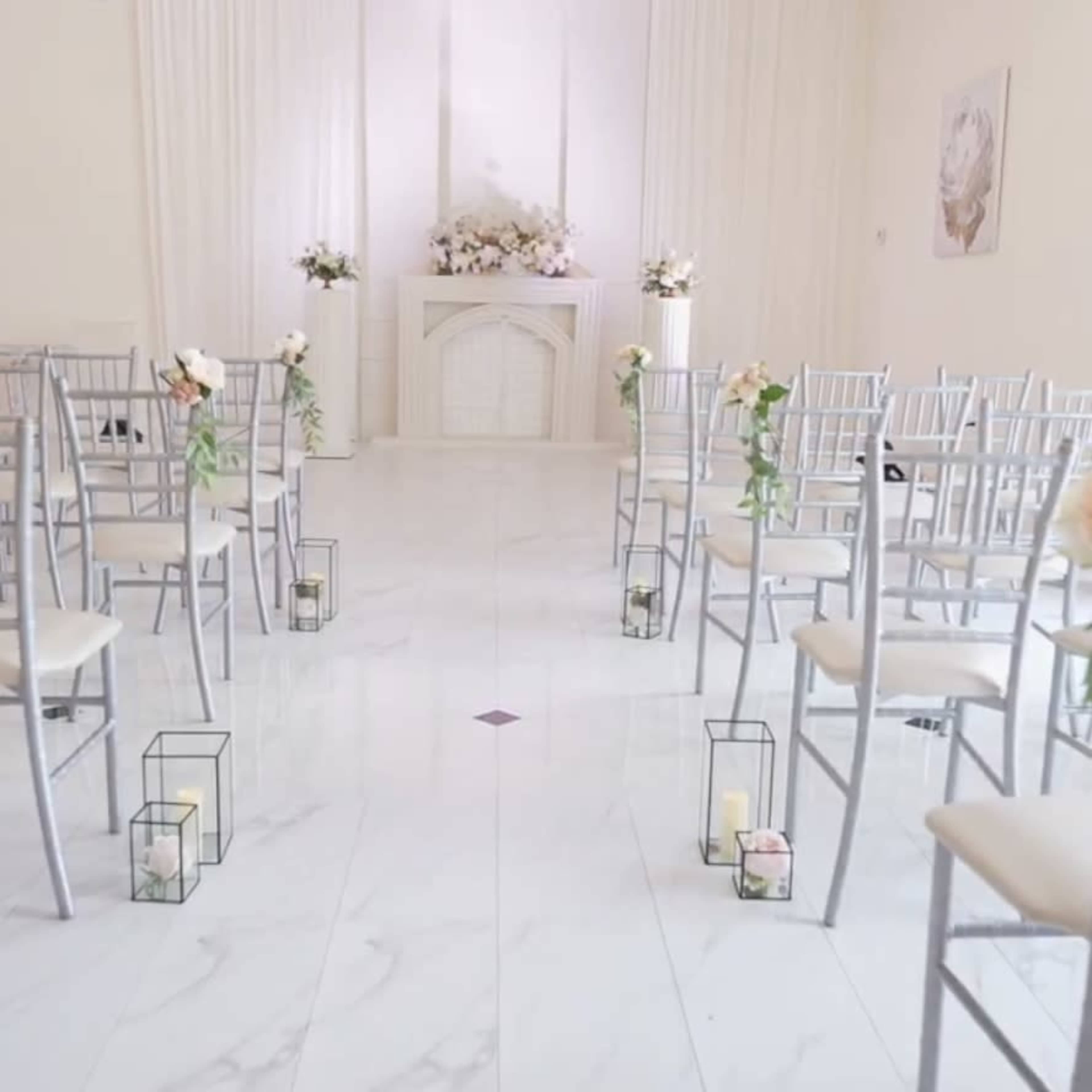 The image depicts a wedding ceremony setup with rows of silver chairs facing a floral-decorated altar in a spacious, well-lit hall.