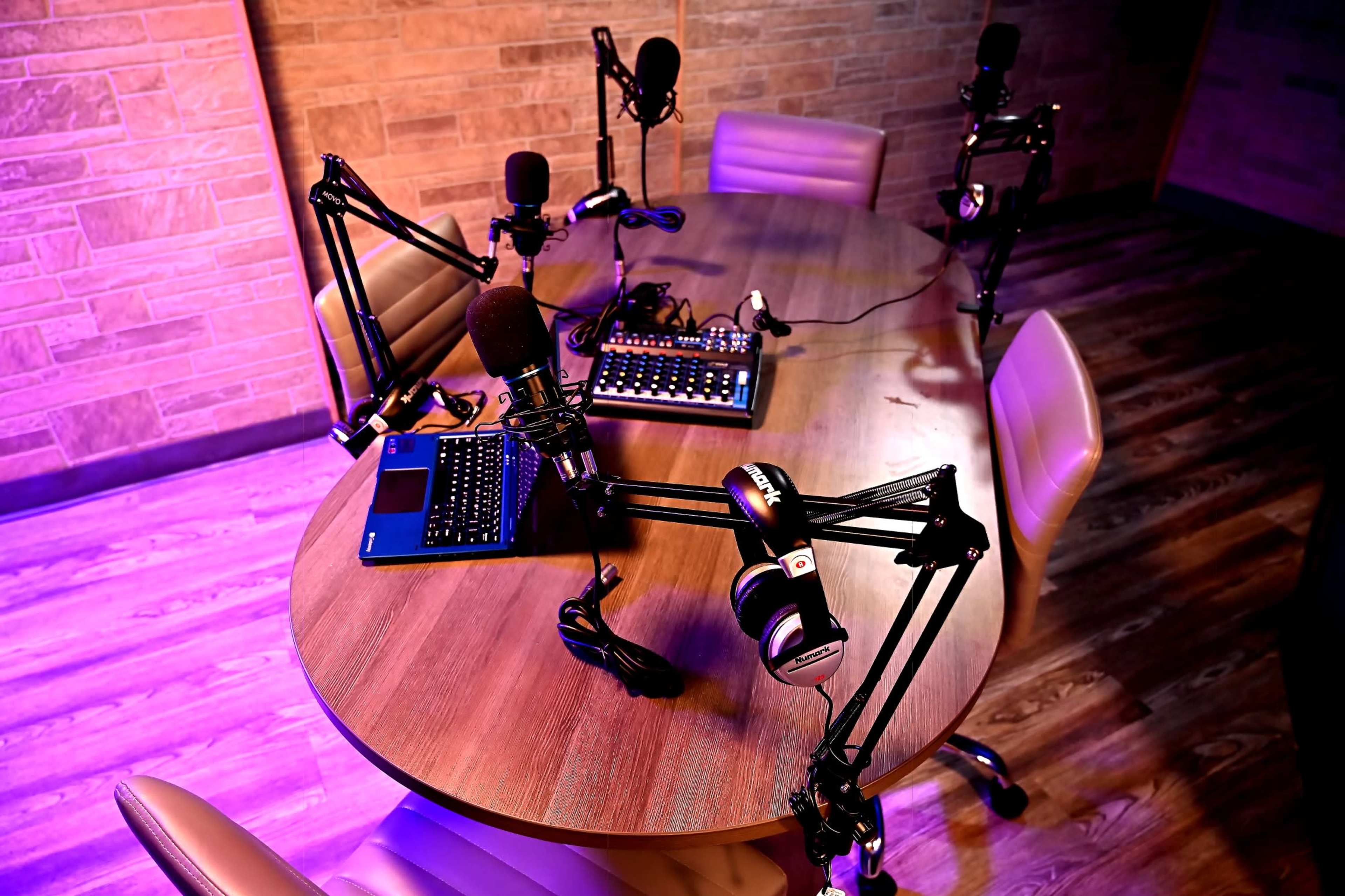 A round wooden table is equipped with microphones, headphones, and laptops in a recording studio setup.