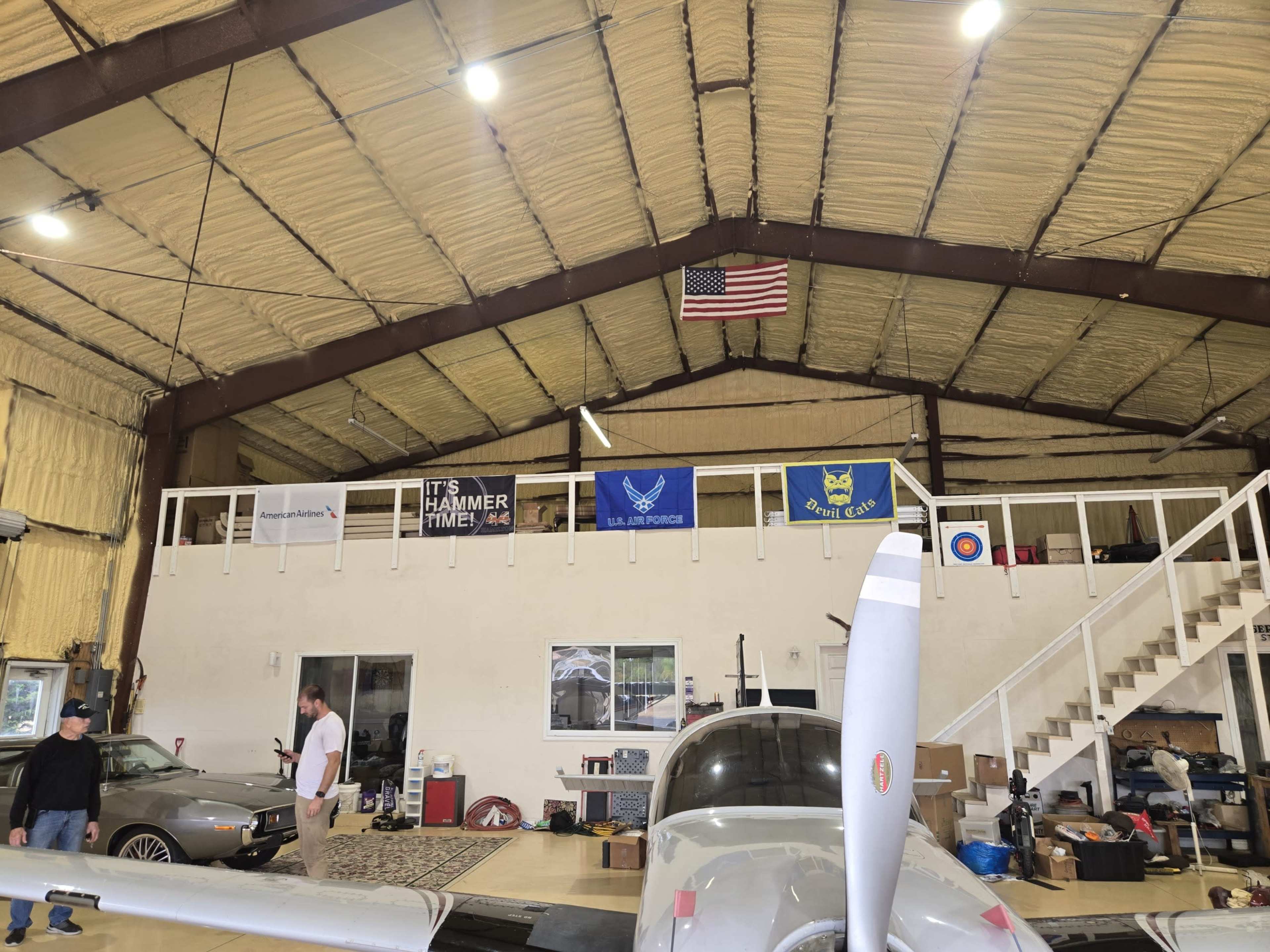 The image shows a spacious hangar with a small aircraft in the foreground, various banners on the wall, and a car parked nearby.