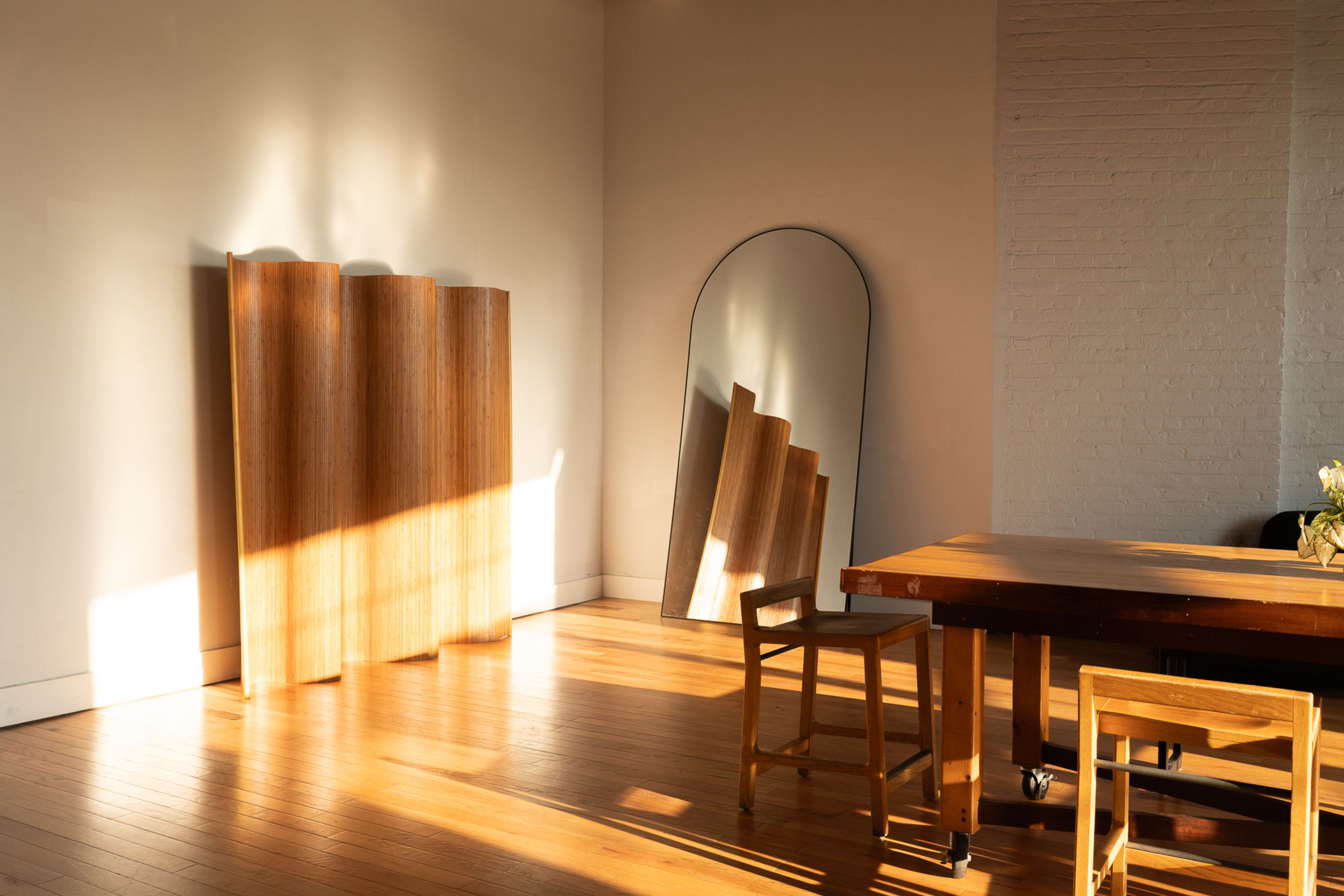 The image features a sunlit room with a wooden table and chairs, a large mirror, and a series of wooden panels leaning against the wall.