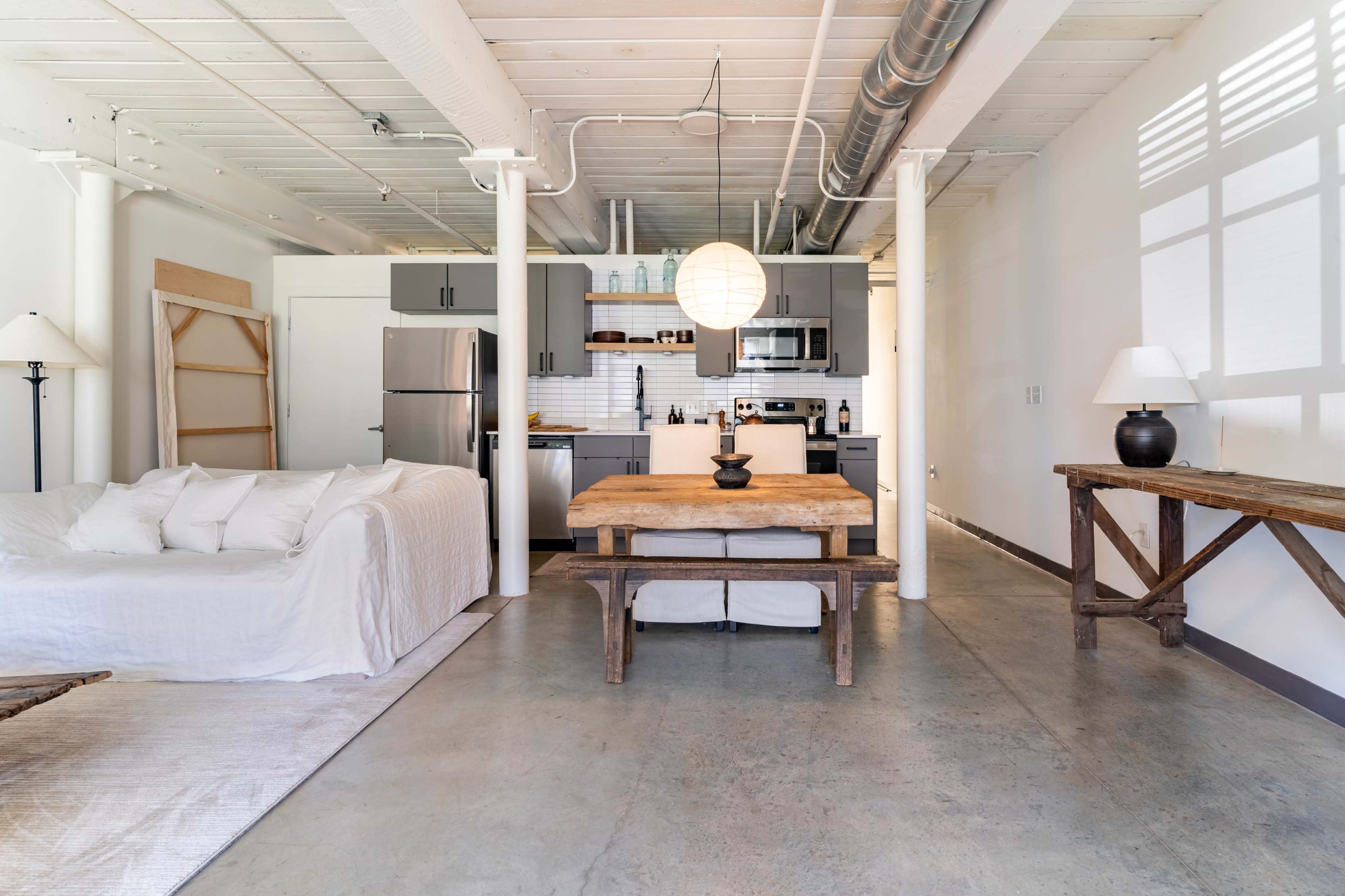 The image shows a modern, open-concept living space with a kitchen, a dining table, and a sofa, featuring minimalist decor and concrete flooring.