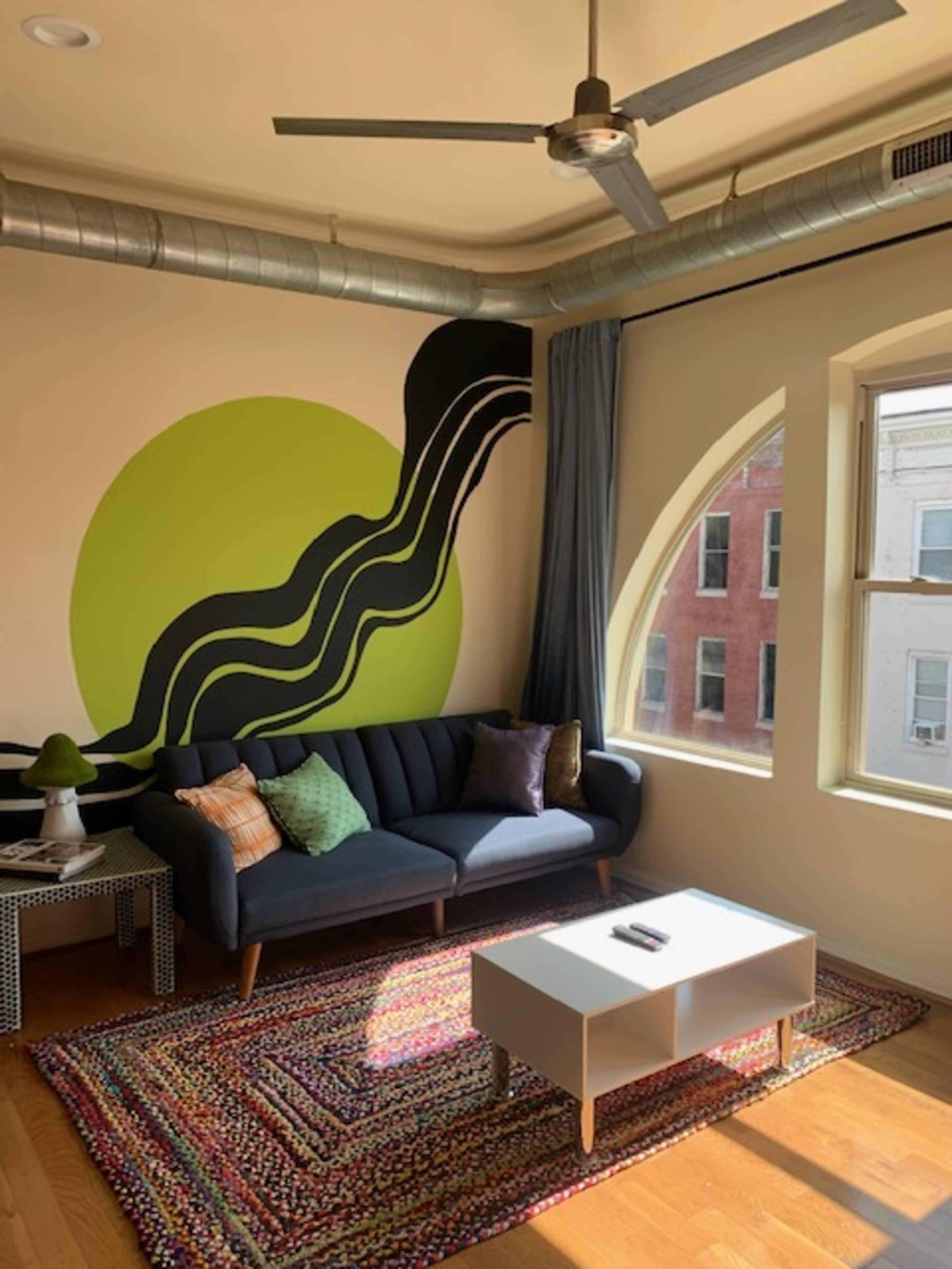 The image shows a modern living room with a dark sofa, a colorful area rug, and a large wall mural featuring bold green and black shapes.