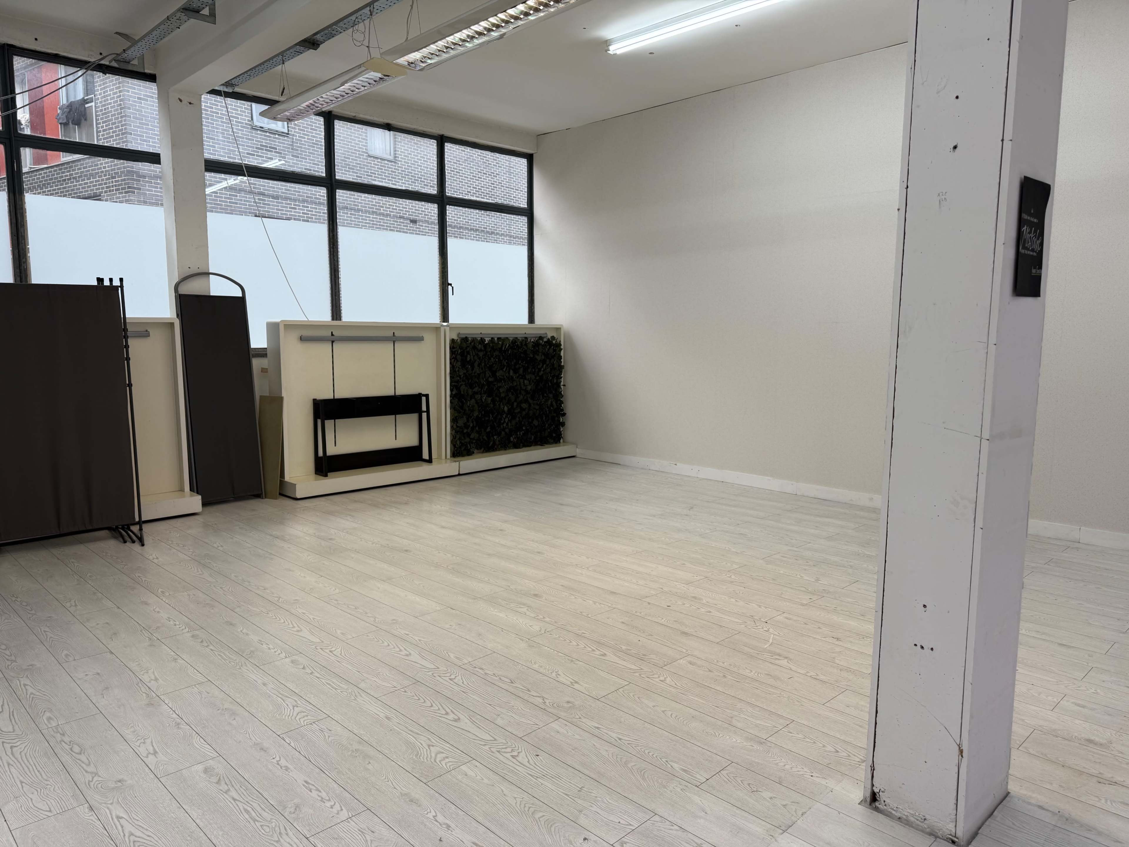 The image shows a vacant room with light flooring, large windows, and a small green feature wall.