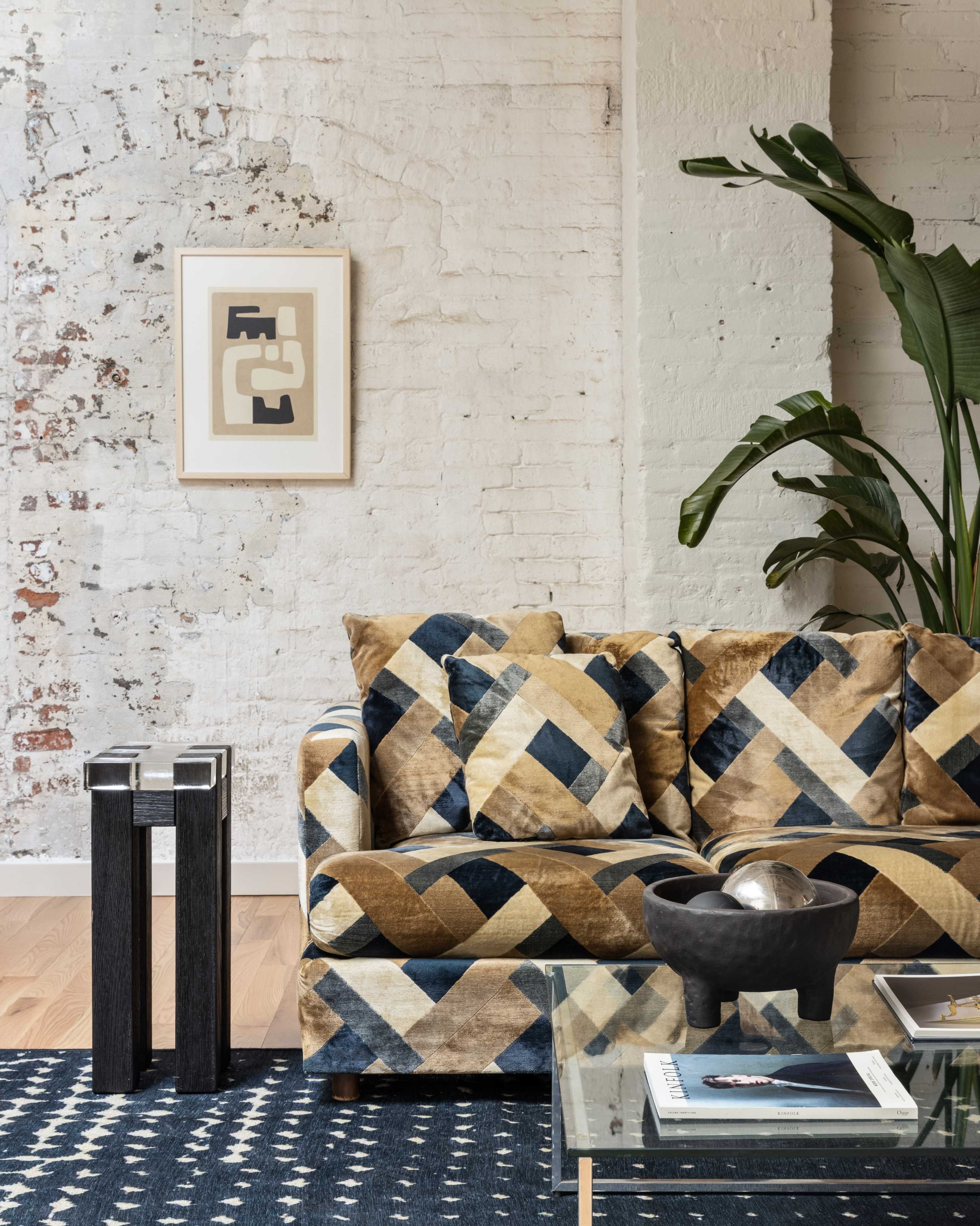 The image depicts a modern living room featuring a patterned couch, a glass coffee table, a decorative bowl, and a vertical plant beside a textured white-brick wall.