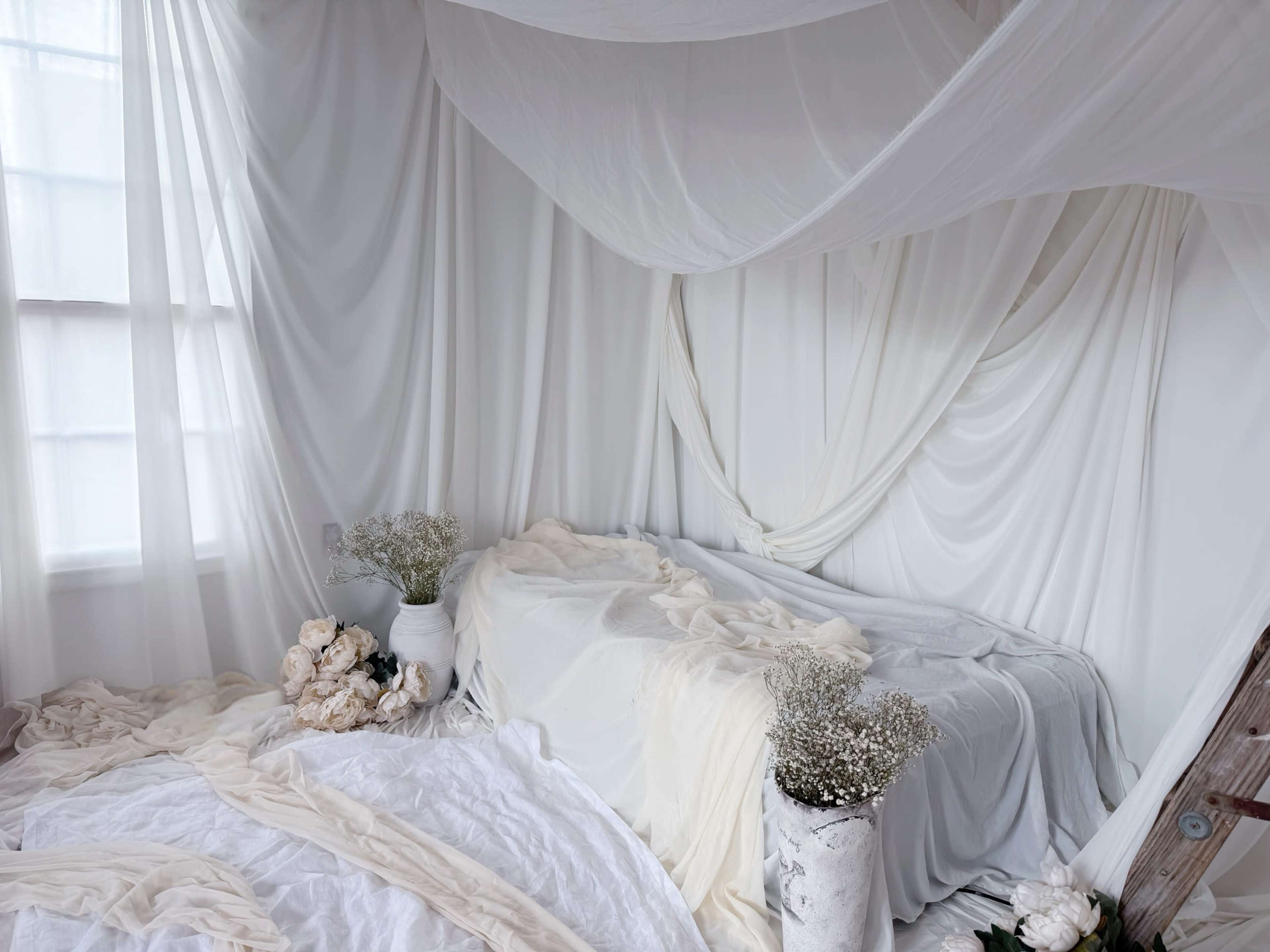 The image shows a softly lit room adorned with flowing white fabrics draped on the walls, a bed covered in layers of white bedding, and potted flowers arranged nearby.