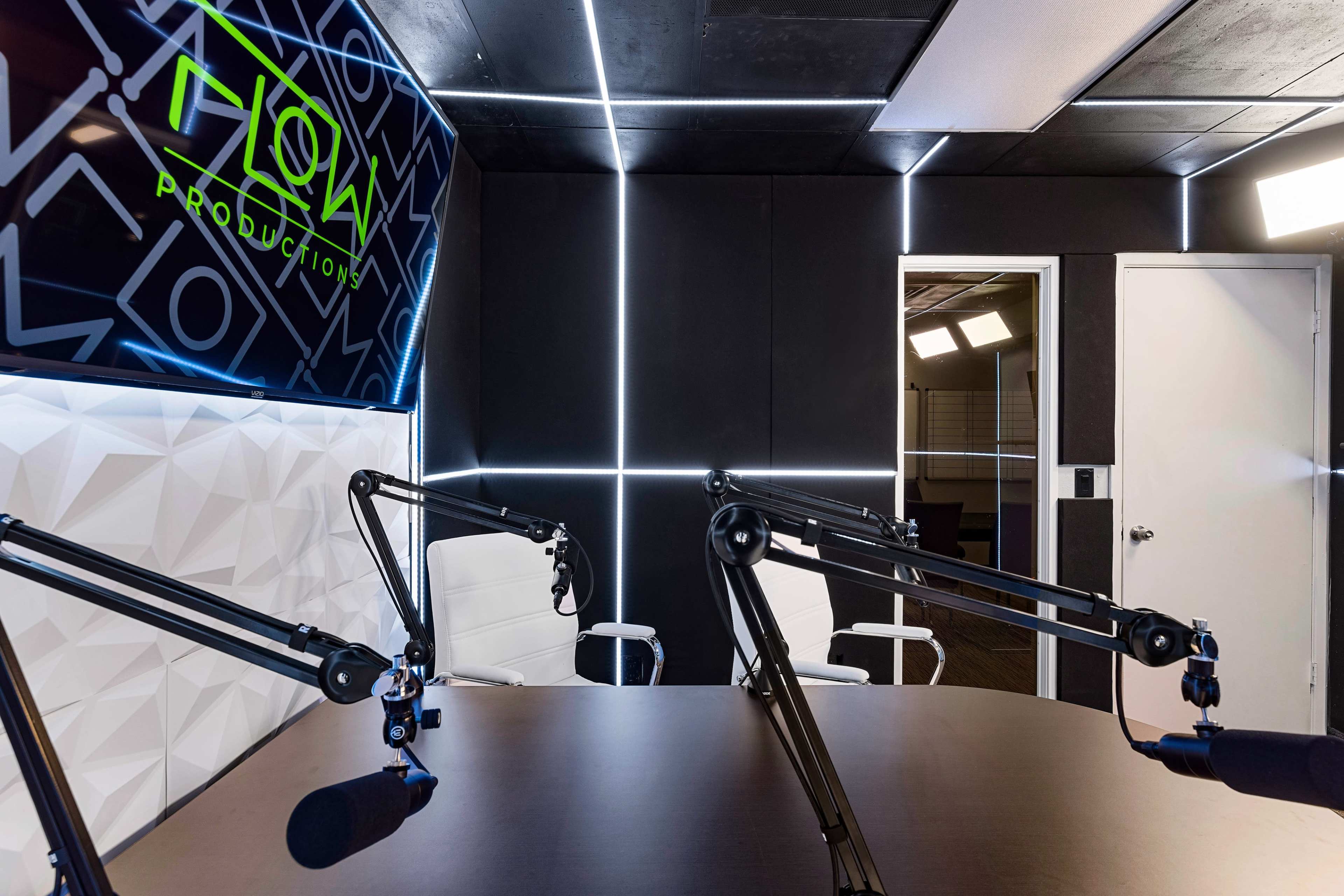 A modern podcast studio with a large branded wall, two adjustable microphone arms, and a round table surrounded by white chairs.