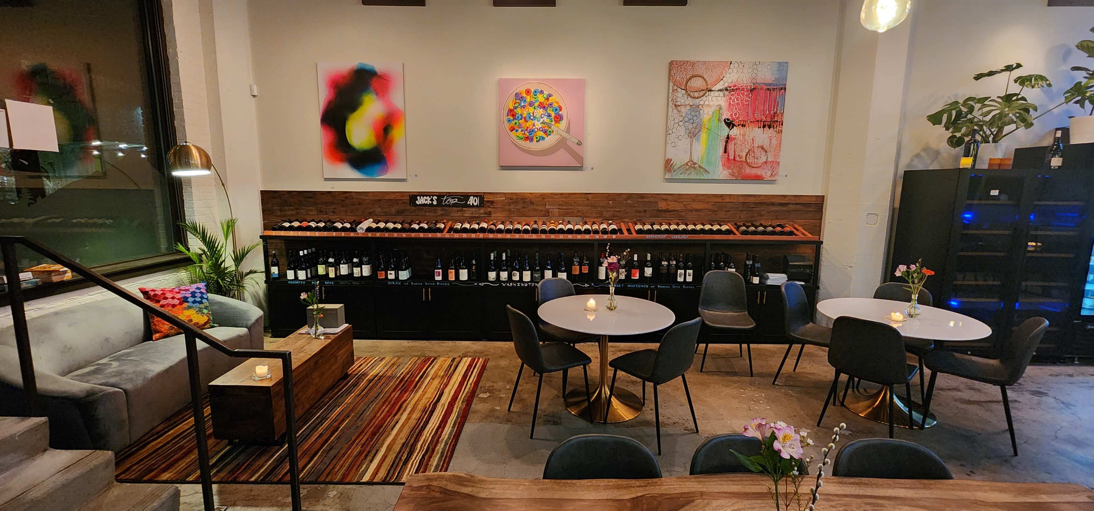 The image shows a modern wine bar featuring circular tables, a dark wooden counter displaying wine bottles, and colorful artwork on the walls.